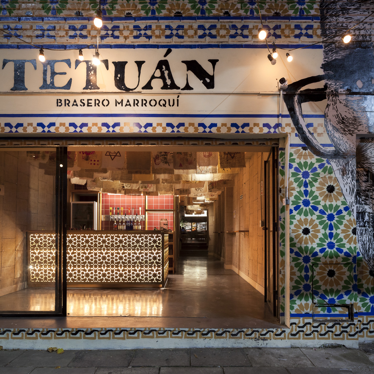 Restaurant entrance with patterned tile frieze, hand-painted lettering and a backlit perforated bar counter glowing at dusk