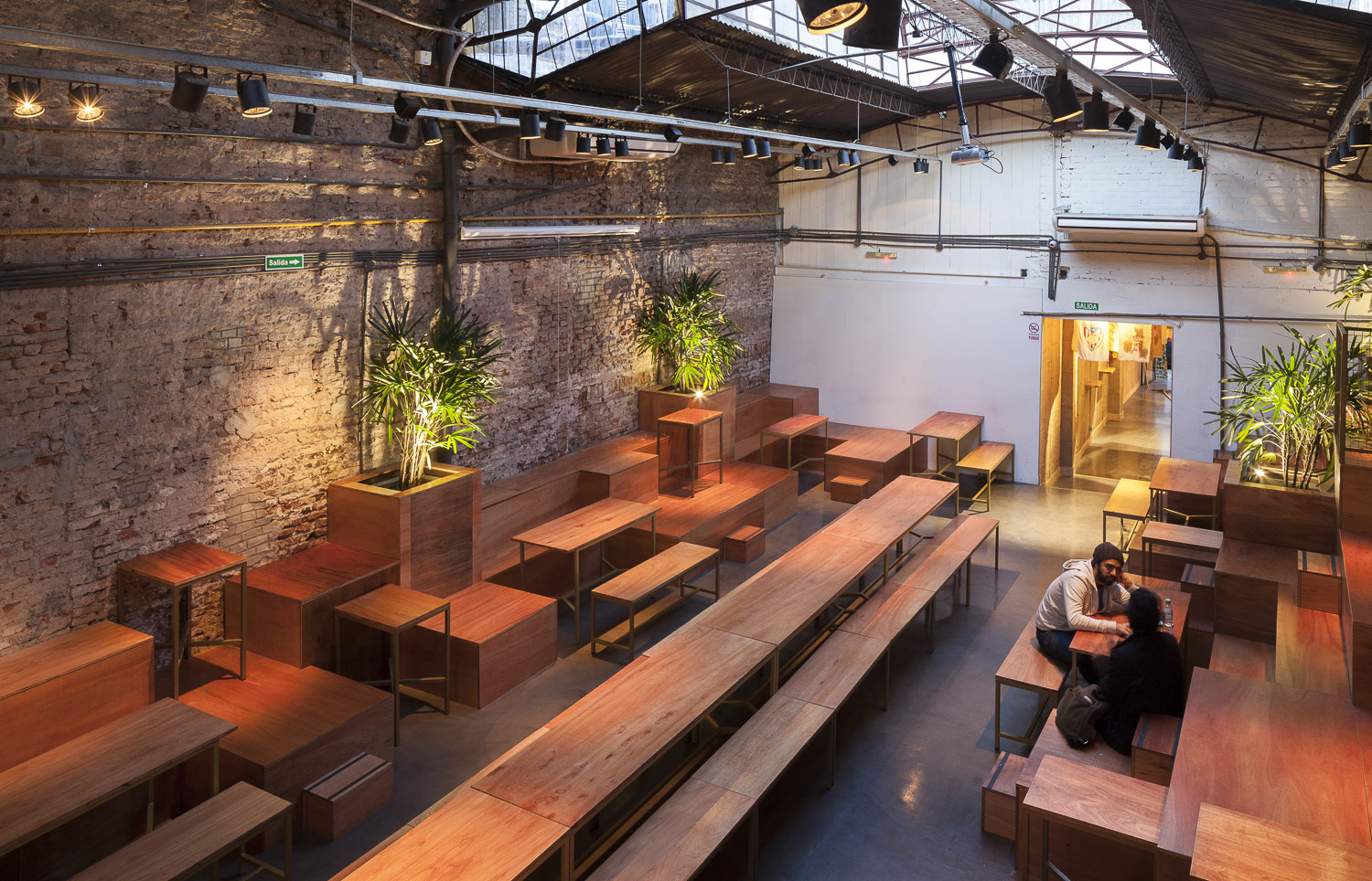High view of long timber tables and stepped seating below a skylit roof, with two diners at the right