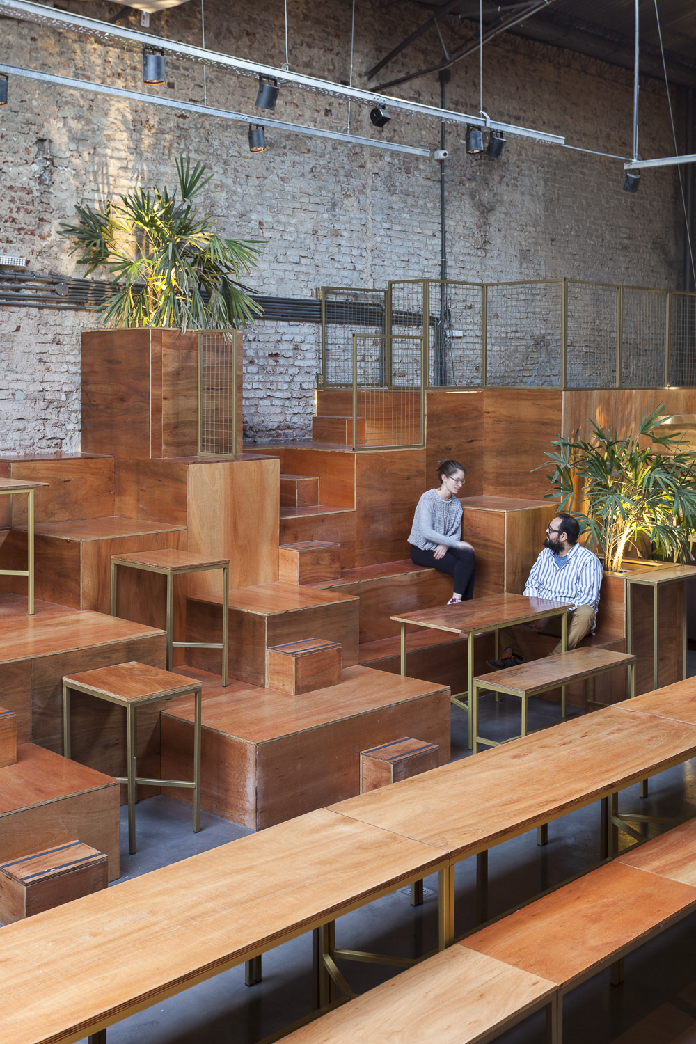 Two people seated on the upper tier of stepped timber seating, with long tables in the foreground and brick wall behind
