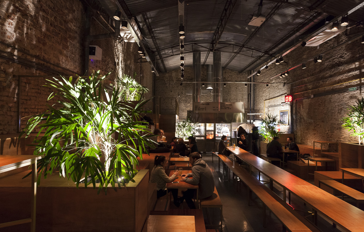 Evening dining hall with diners at long wooden tables, a large lit palm in the foreground and a steel-trussed roof overhead