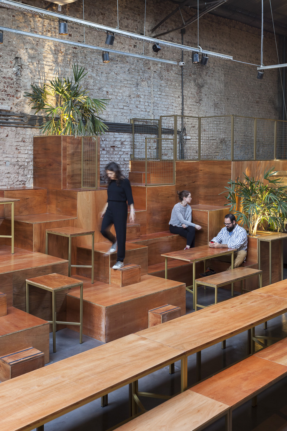 Stepped timber platforms with brass-framed stools and potted palms against an exposed brick wall, a woman descending in daylight