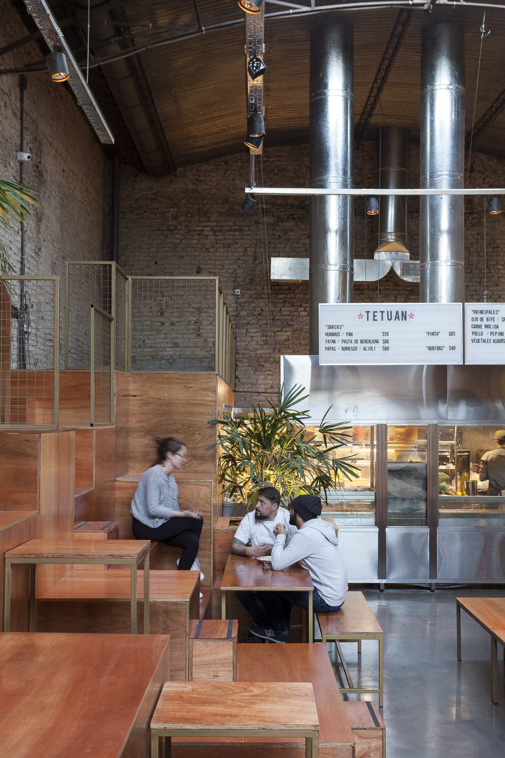 Diners seated at wooden tables in front of a stainless steel open kitchen and twin ducts under a vaulted roof