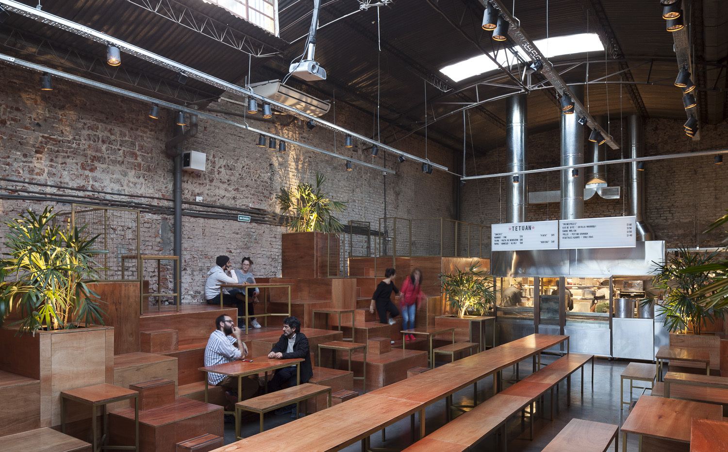 Wide view of the tiered timber seating, long communal tables and steel kitchen hood beneath skylights in a brick hall