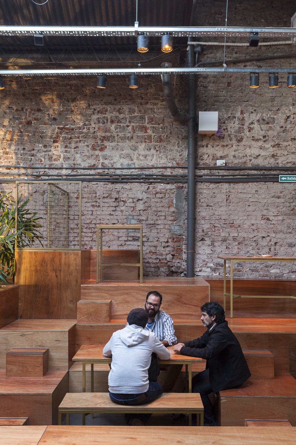 Three men talking at a small table on timber steps, with a brick wall and spotlight rail behind