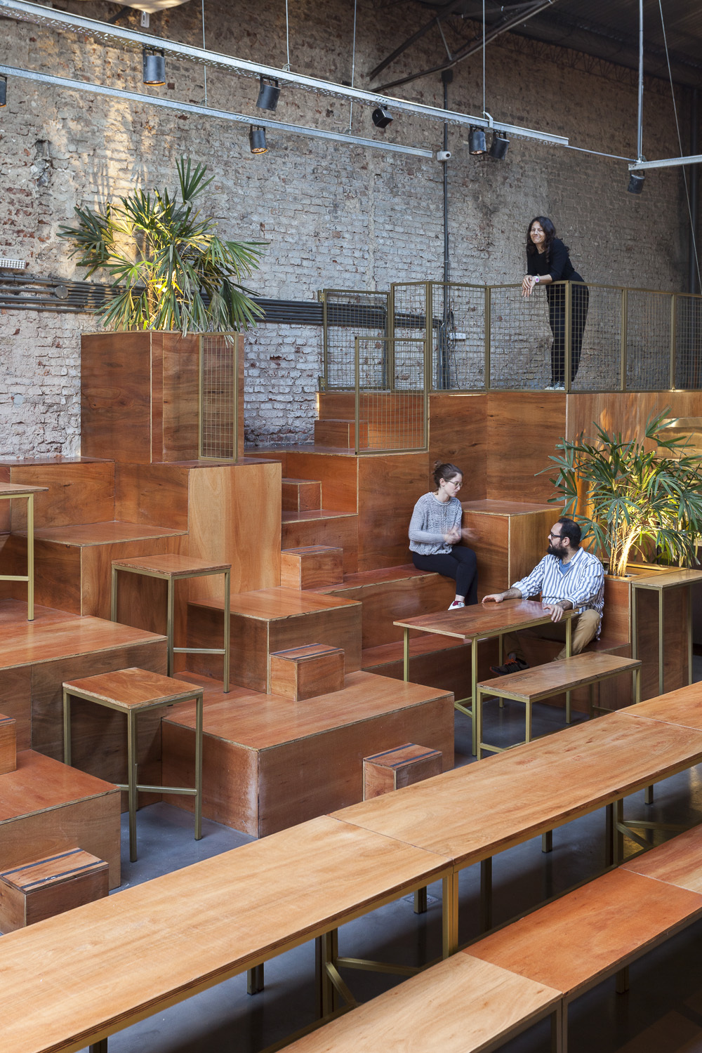 Woman leaning on the mesh railing above stepped timber seating while two people sit at a table below