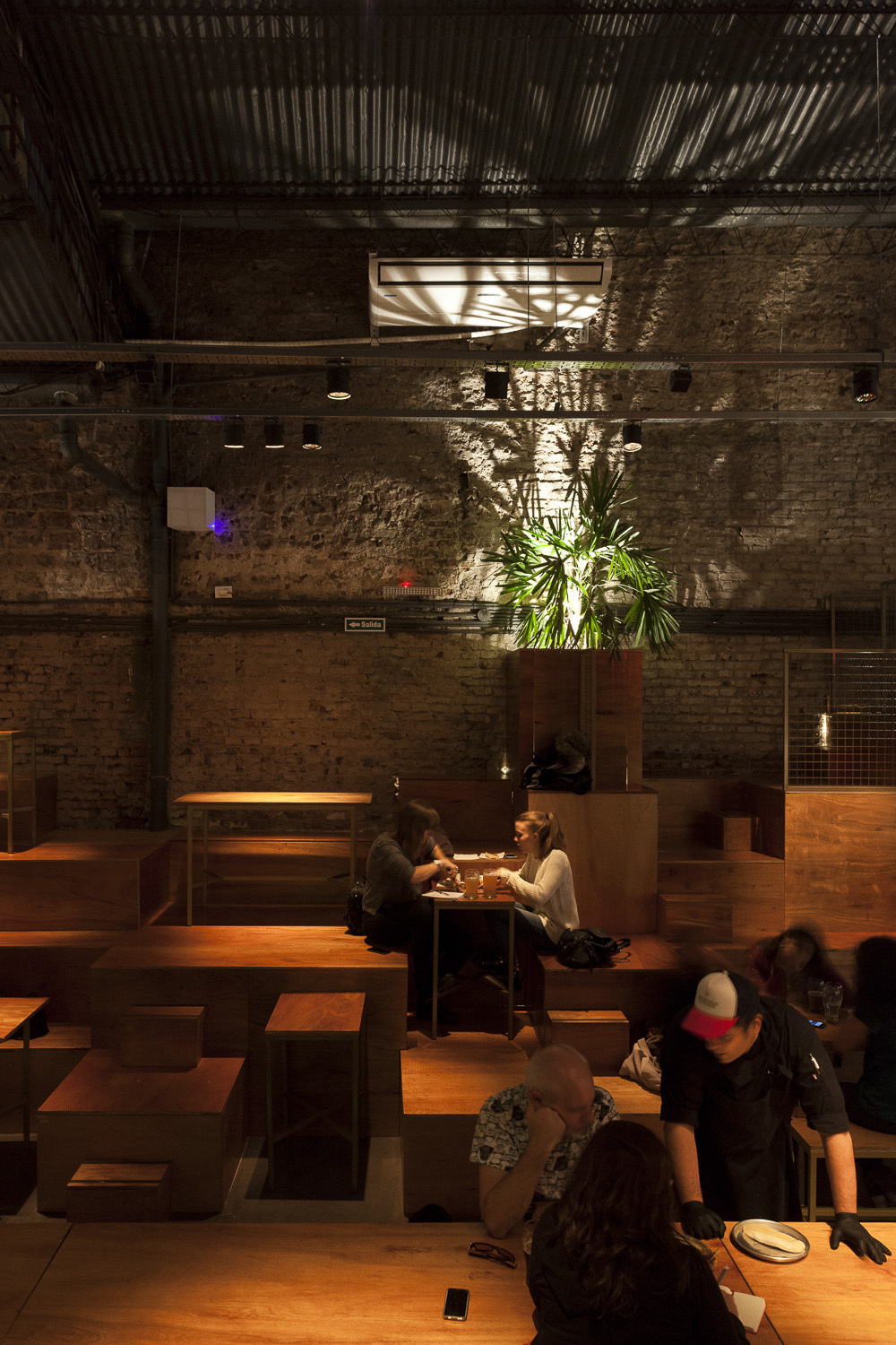 Evening view of timber tiers and diners, a cook in a red cap, and dappled light across a brick wall