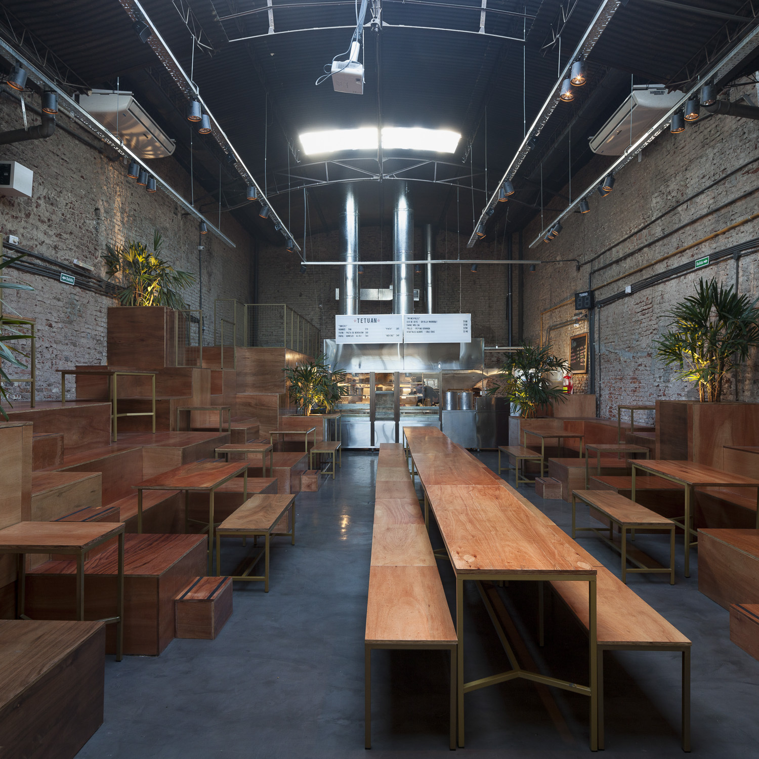 Long dining hall with stepped timber platforms, brass-framed tables and benches, potted palms and exposed brick walls lit by a bright skylight