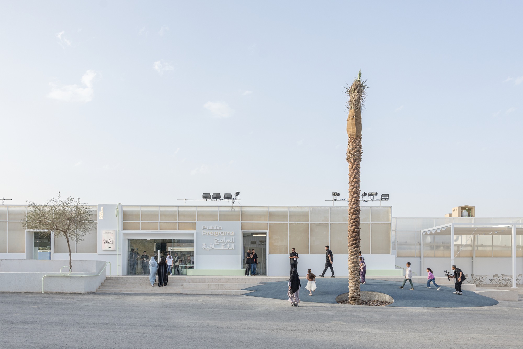 Entrance facade of translucent panels with a single tall palm trunk rising from a circular paved forecourt, visitors and children crossing in daylight