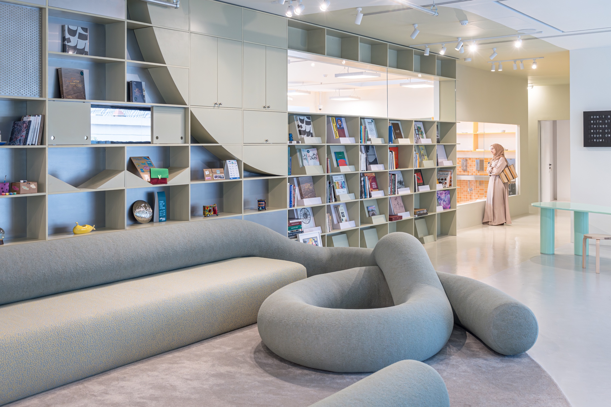 Reading lounge with a looping grey upholstered sofa in front of sage shelving filled with books, a visitor browsing at the back