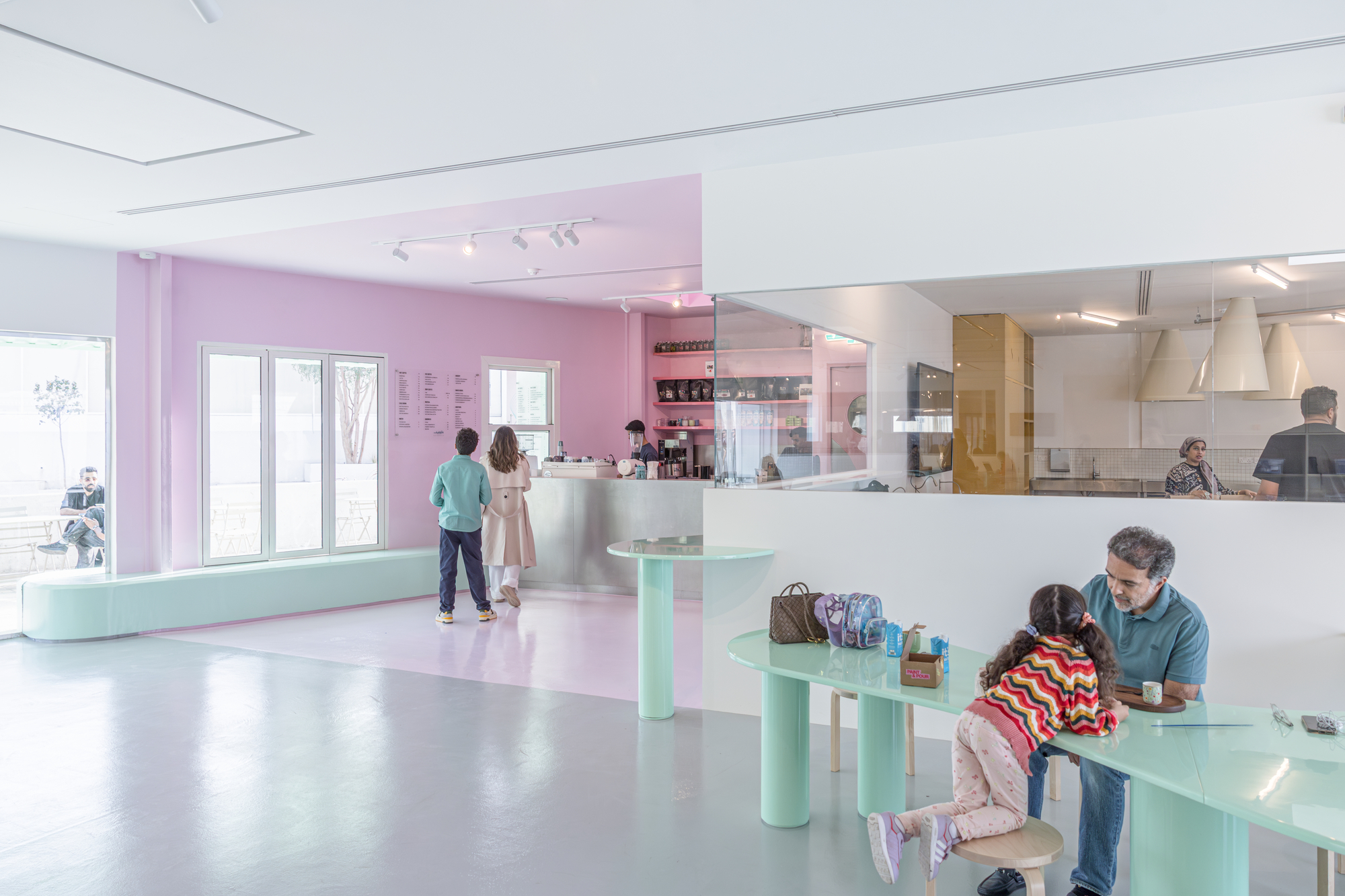 Pink-walled cafe with mint green tables and a bench, a child and adult seated at the counter beside the open kitchen