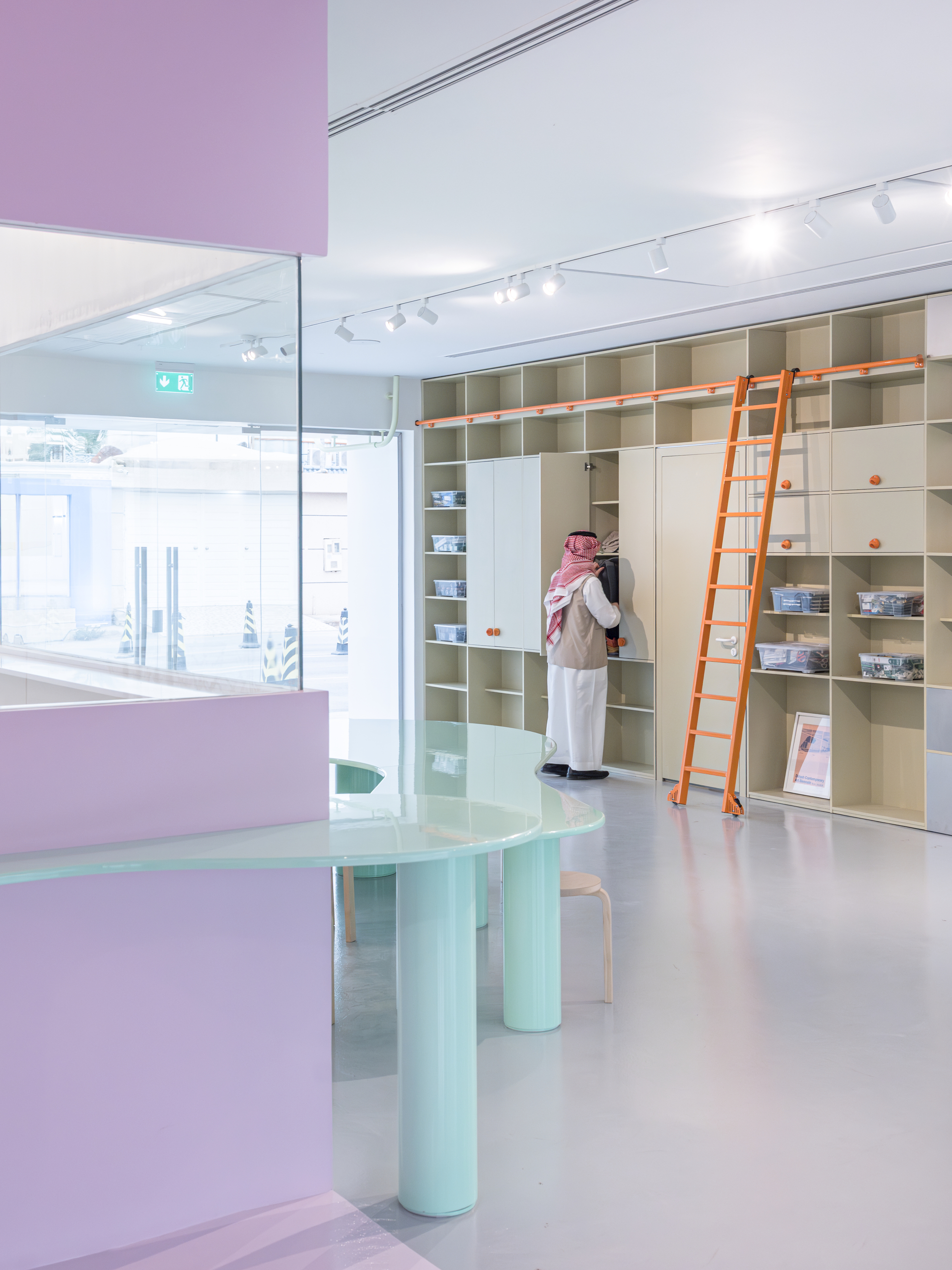 Pale shelving wall with an orange rolling ladder, a man in traditional dress reaching into a cabinet beside a mint table