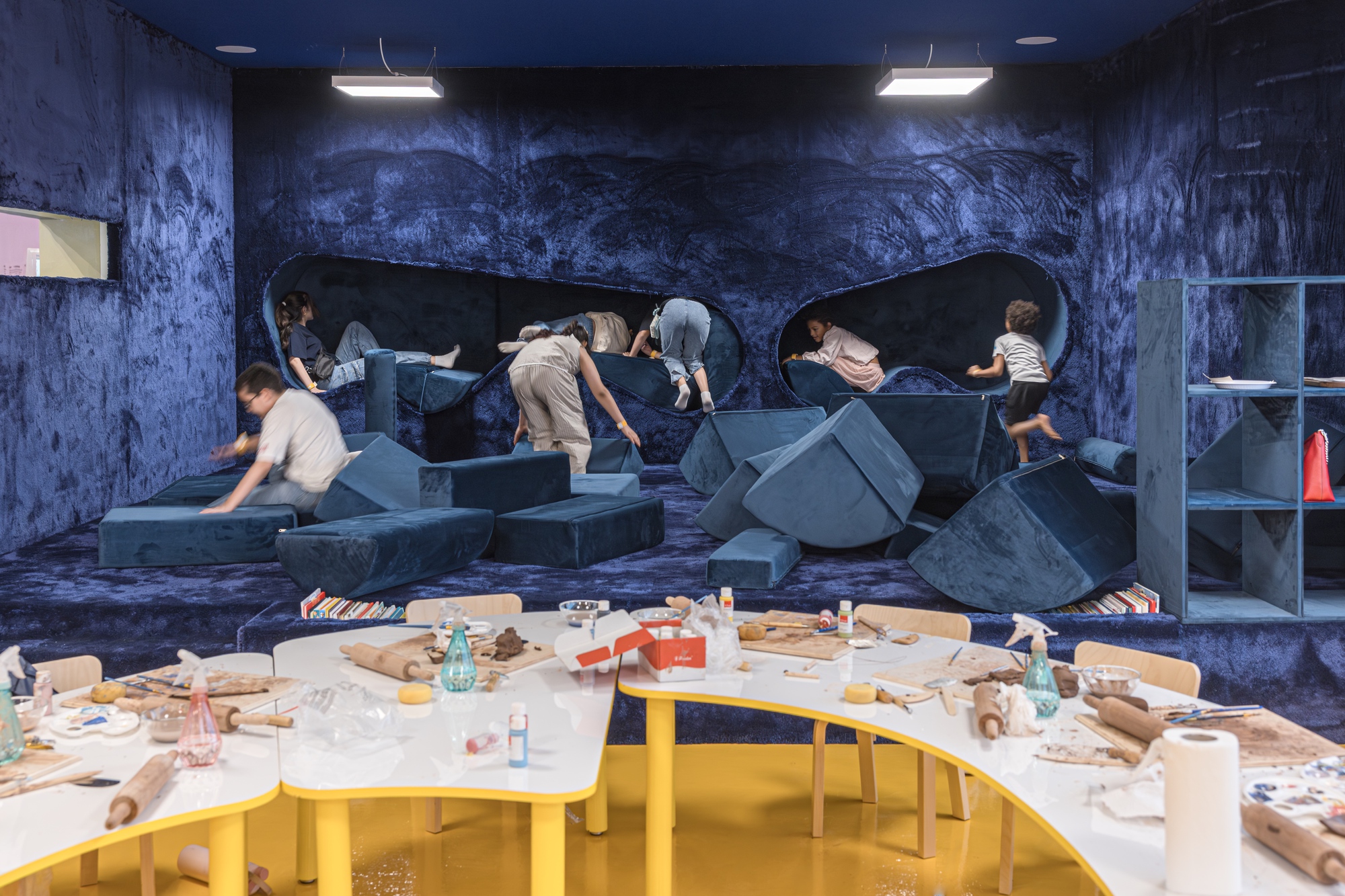Children climbing over blue velvet foam blocks and alcoves above yellow-edged craft tables scattered with rolling pins and clay tools