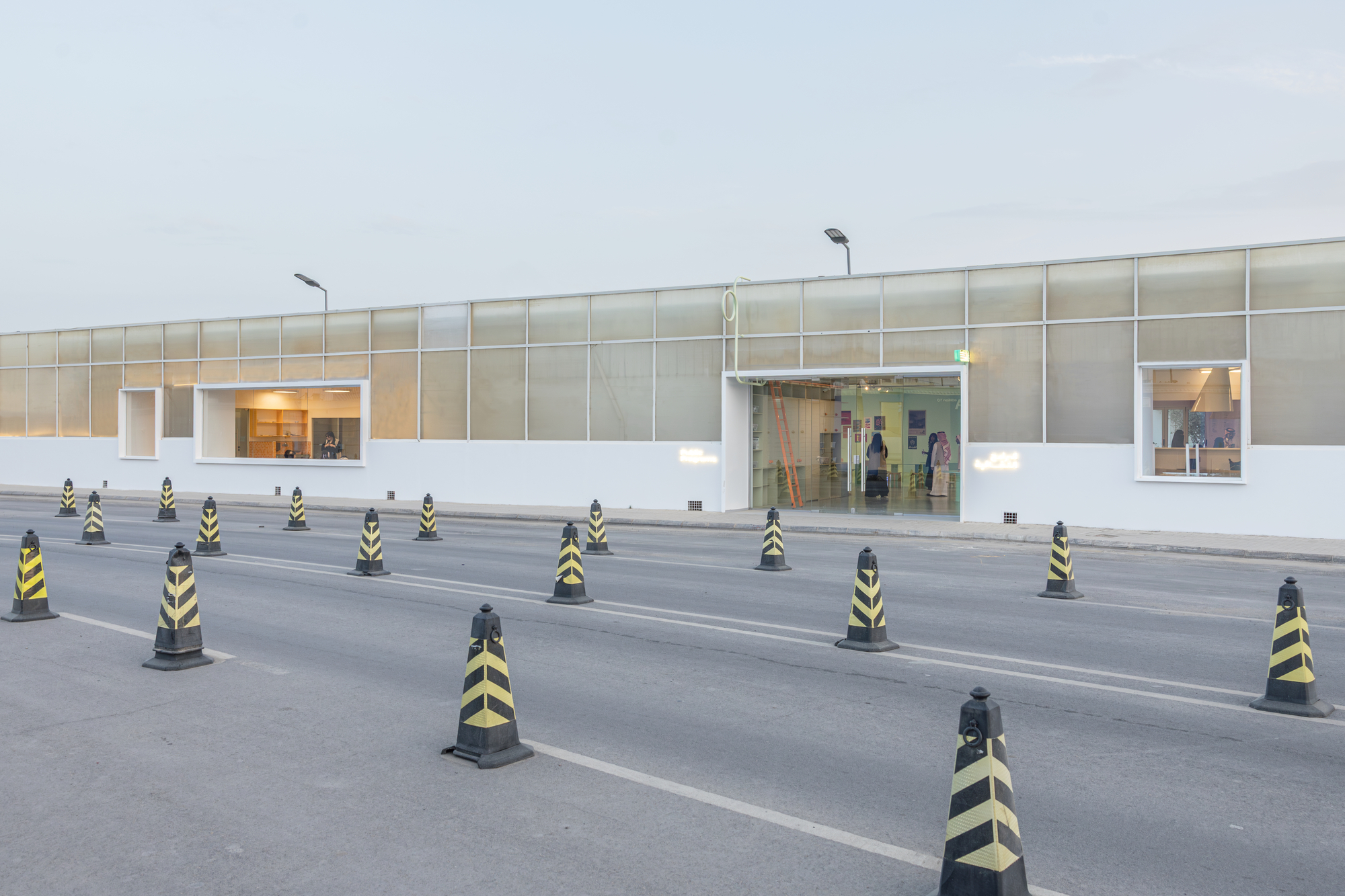 Long translucent street facade with framed openings, lined by black and yellow chevron traffic cones along the empty road at dusk