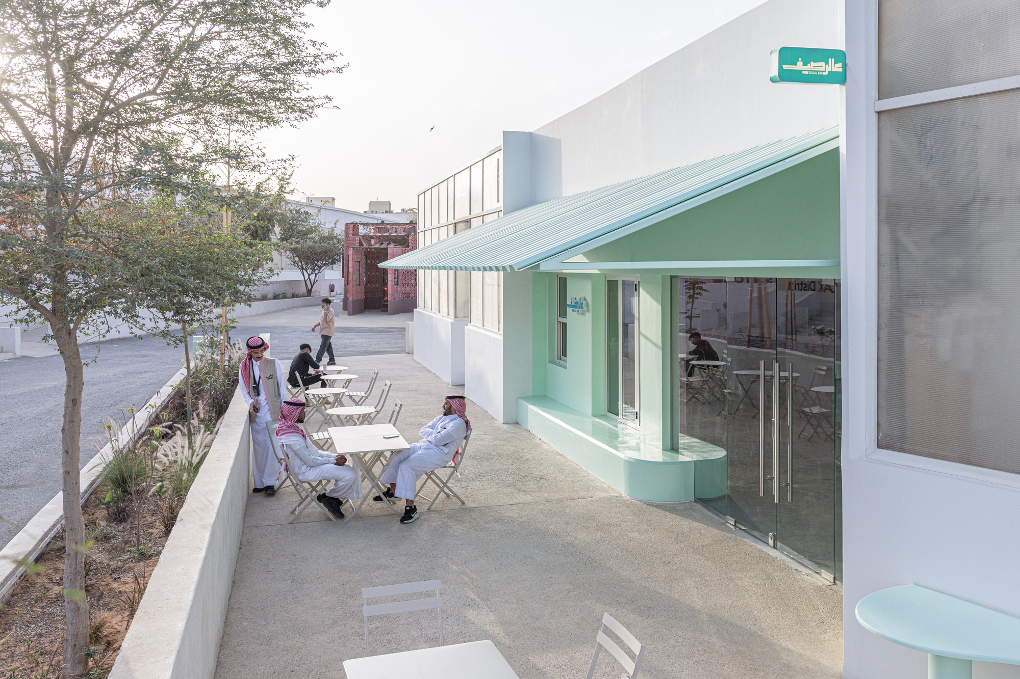 Mint green porch canopy and curved plinth beside a glazed entrance, with men seated at folding tables on a terrace in daylight