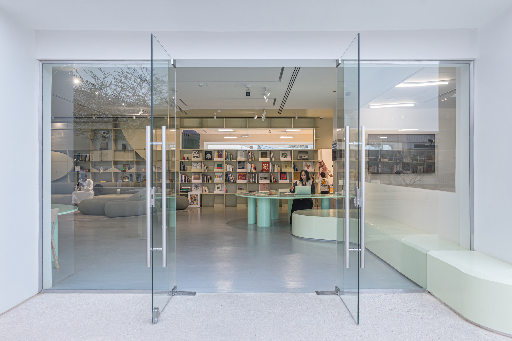 Open pivot glass doors revealing a bookshop with pale shelving, a mint round-legged table and a seated attendant
