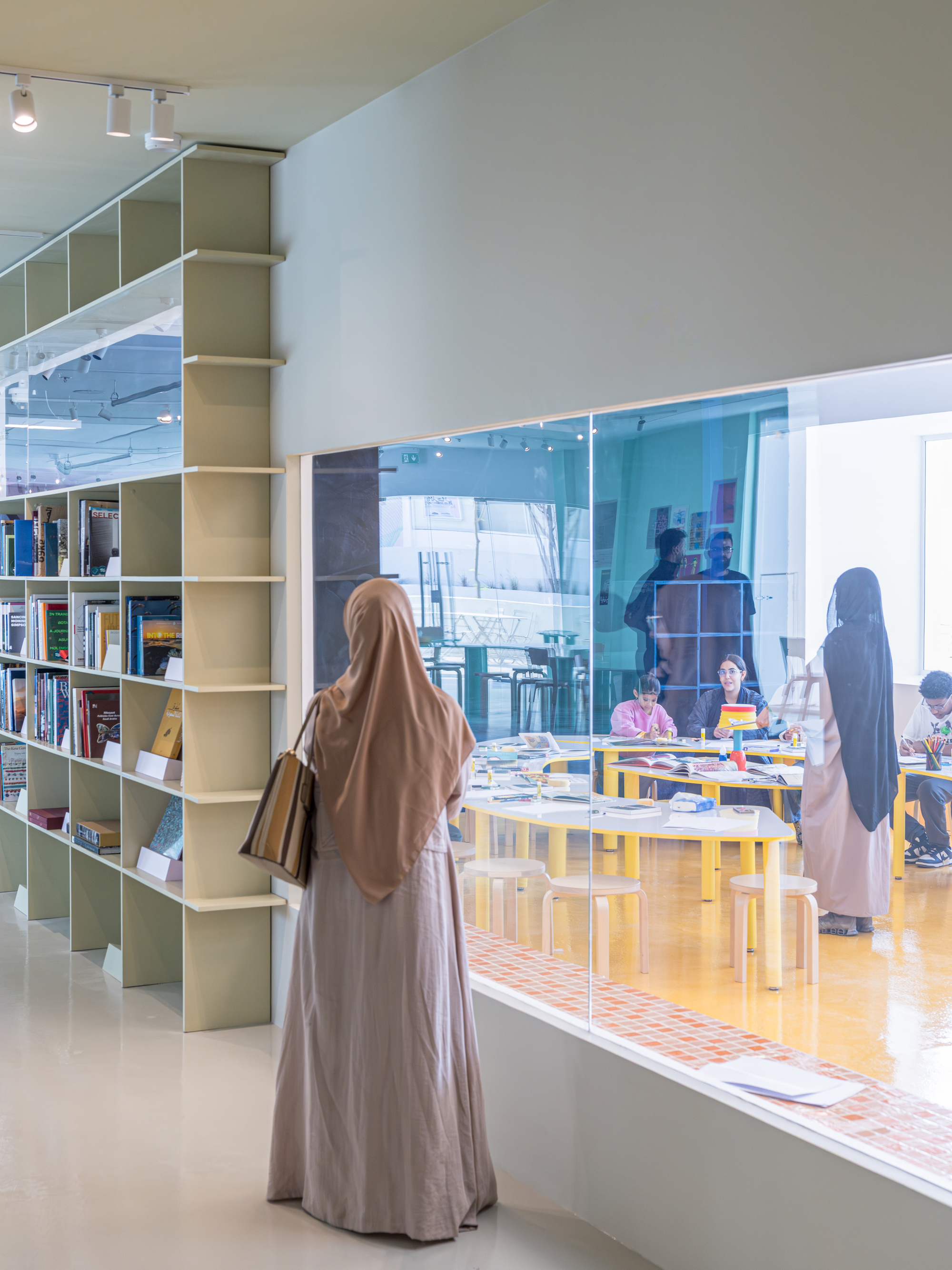 Woman in a headscarf facing a glazed window onto a workshop room with yellow tables, beside olive bookshelves