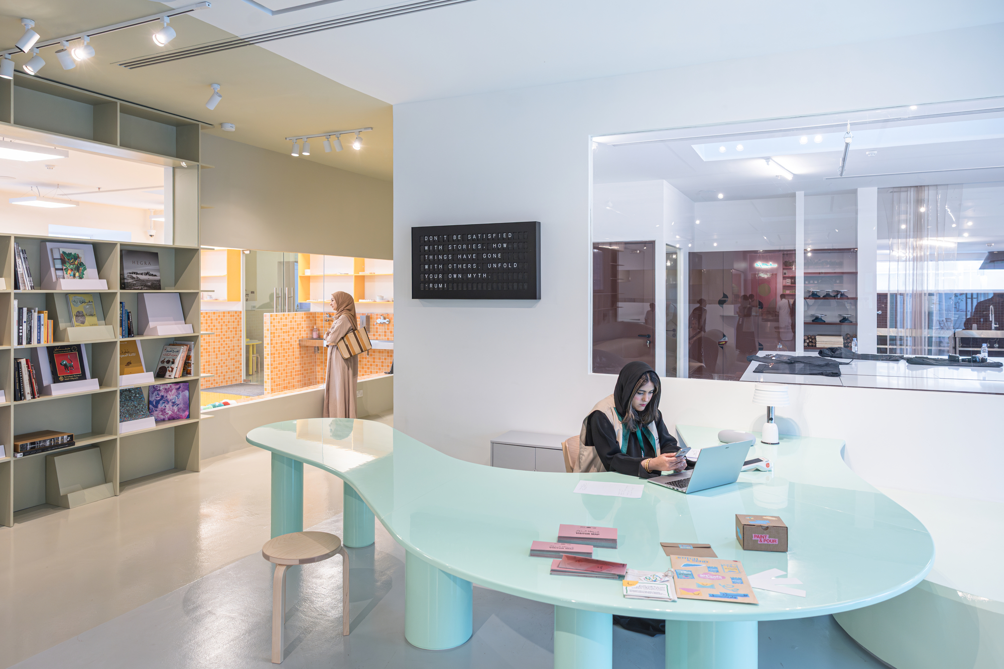 Curving mint green desk with cylindrical legs and a woman at a laptop, with book shelving and a text panel behind