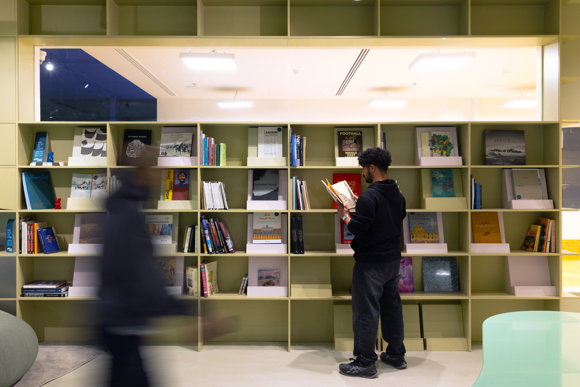 Man browsing olive green display shelves of art books while a blurred passerby moves across the foreground