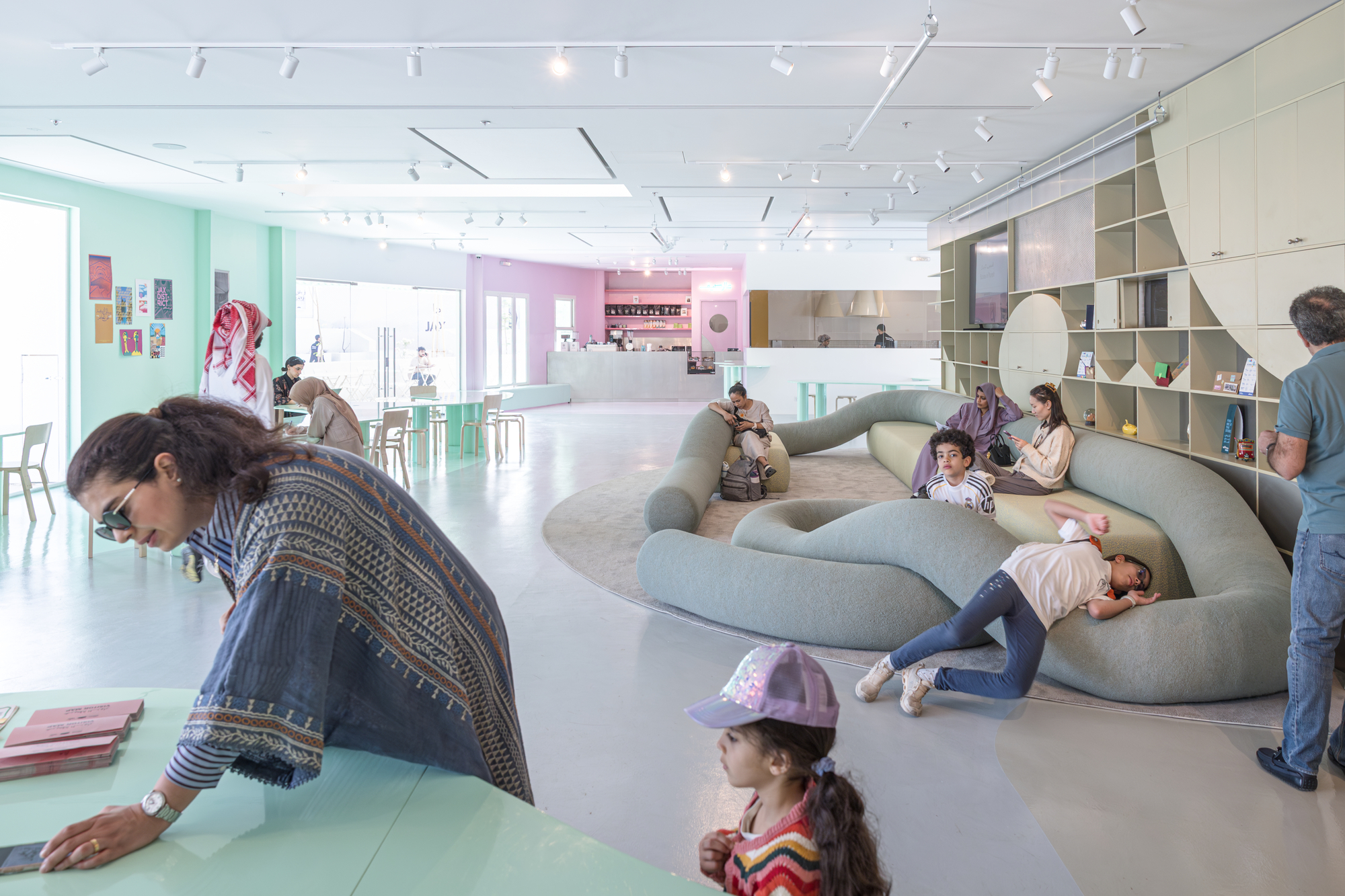 Open reading hall with a sinuous grey-green sofa, children playing on it, and a mint table in the foreground