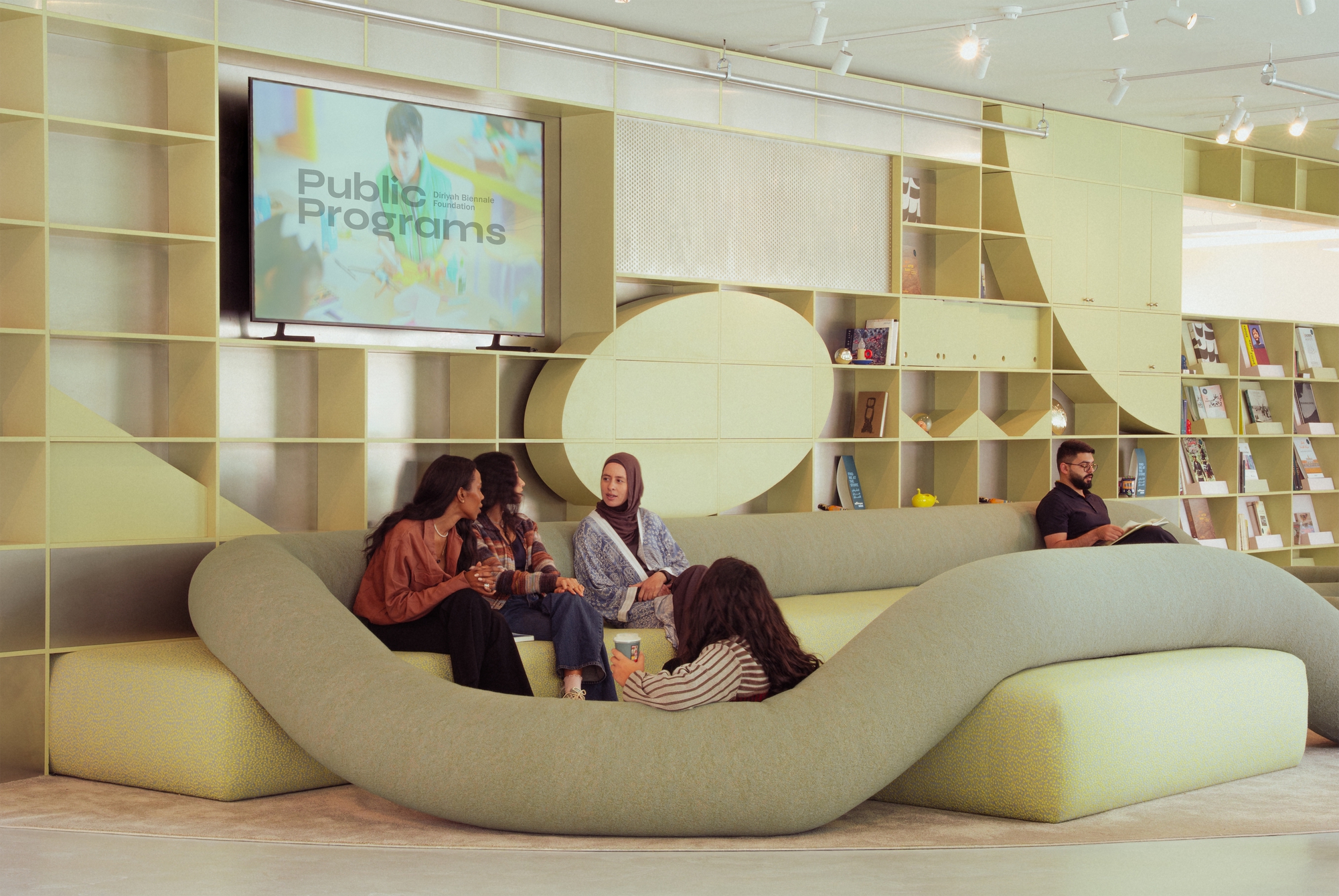 Visitors seated on a curving sage green sofa in front of yellow-green shelving and a wall screen reading Public Programs