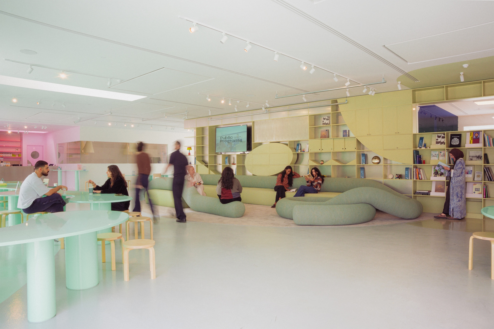 Wide view of a pale green lounge with sinuous seating, round mint tables, wooden stools and blurred figures walking past