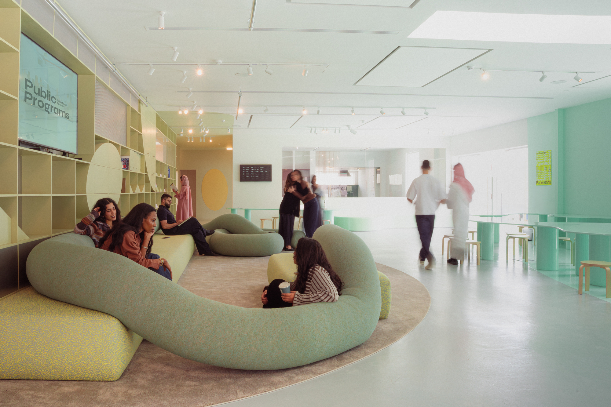 Looping green sofa on a round carpet, with people lounging and a pair walking past mint tables in soft daylight