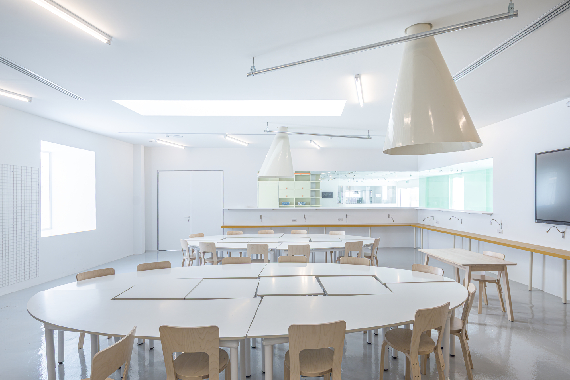 Classroom with white oval tables pushed together, birch chairs, cone-shaped pendant lamps and a skylight washing the room in daylight