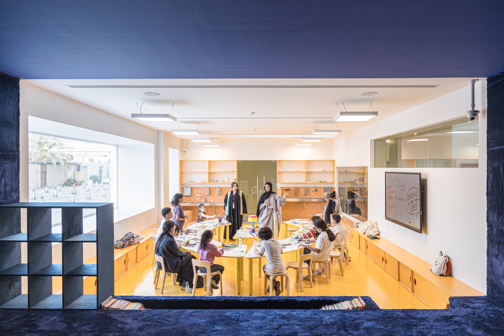 Children seated at yellow-edged tables in a bright classroom, framed by an opening lined with deep blue pile fabric and a cubby shelf