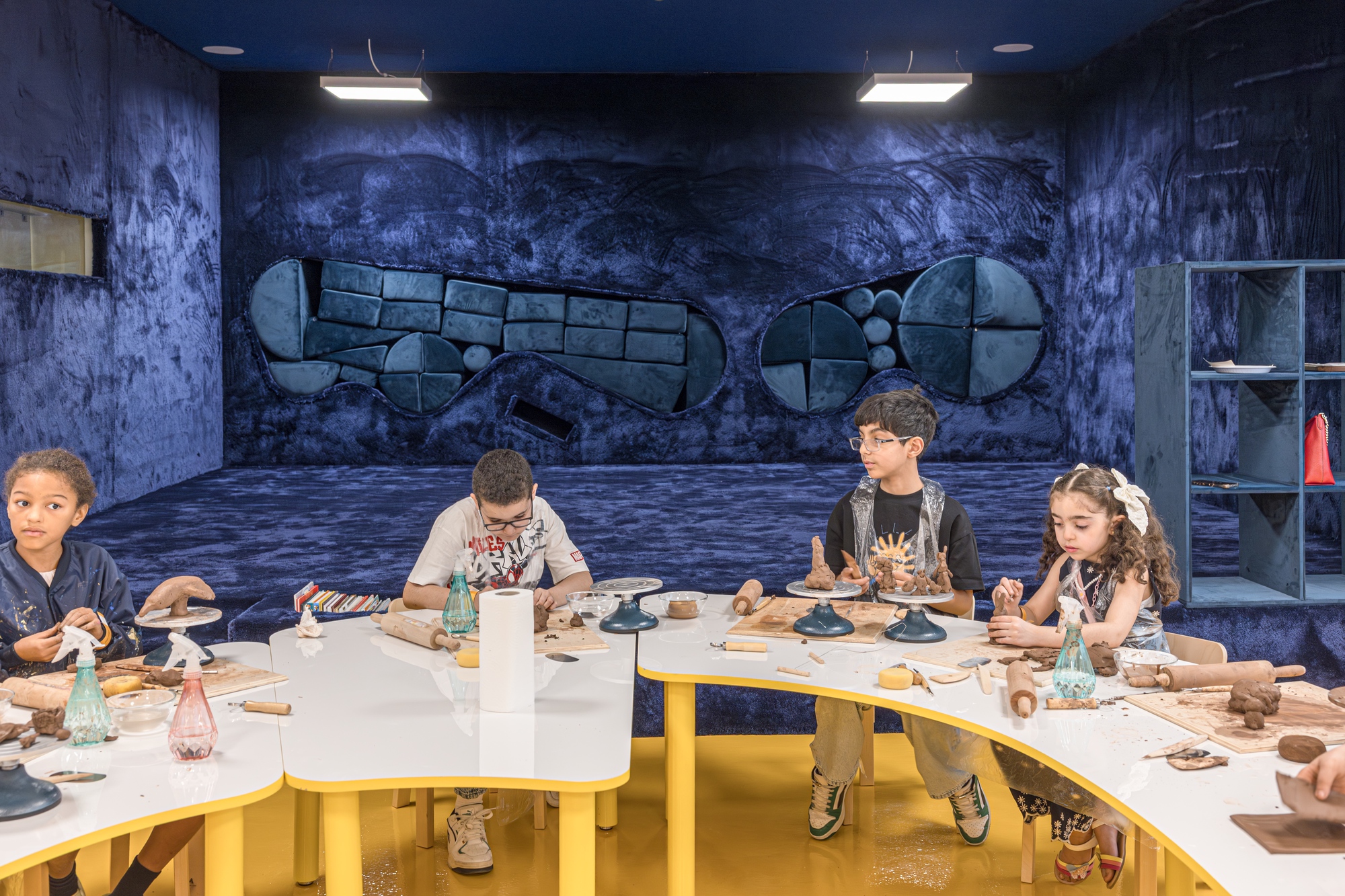 Four children shaping clay at curved white tables with yellow legs in front of a blue velvet wall with carved stone-pattern recesses