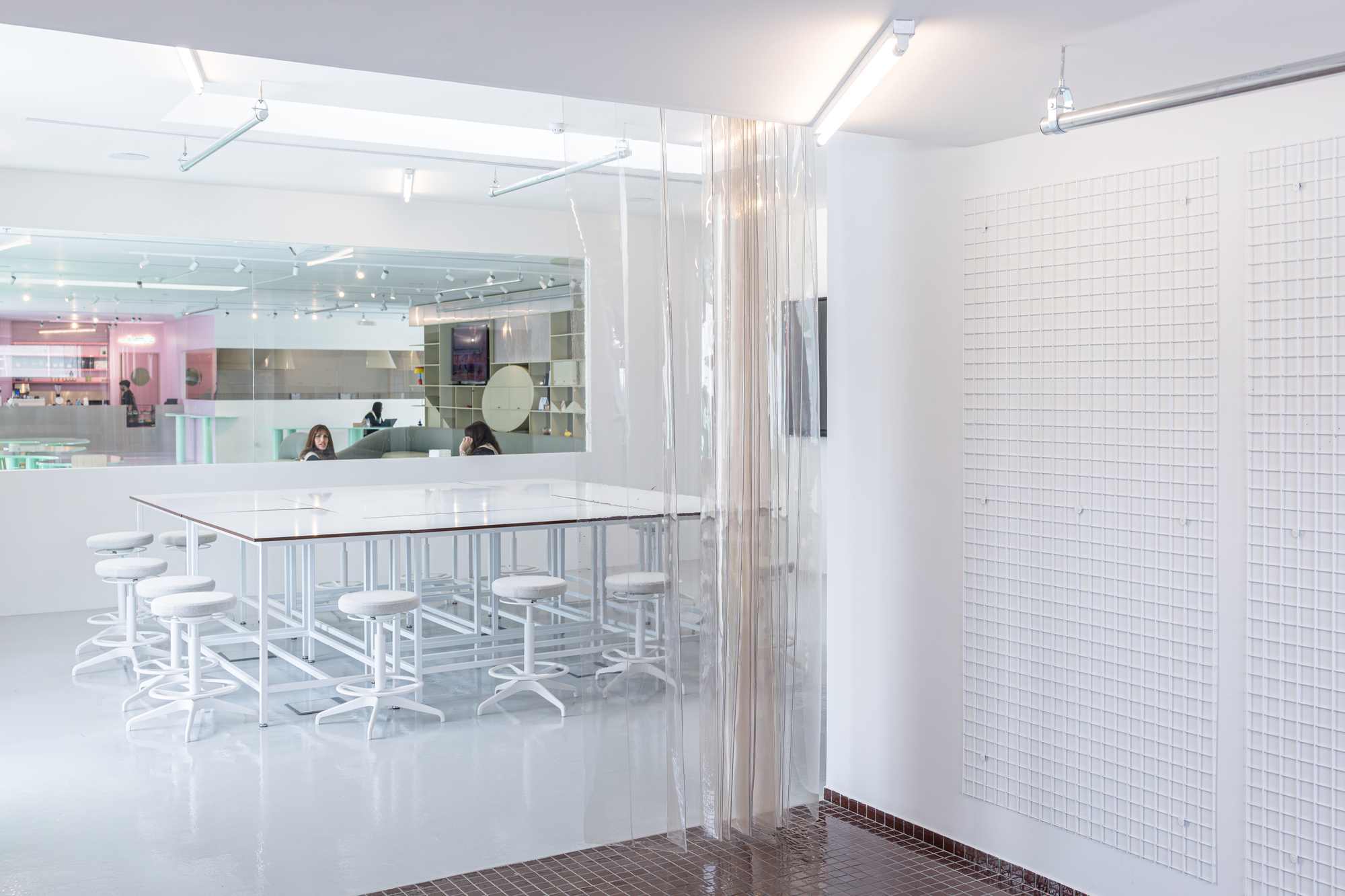 White stools around a glass-topped table beside a translucent plastic strip curtain, with a gridded wall panel and a pink-toned room beyond