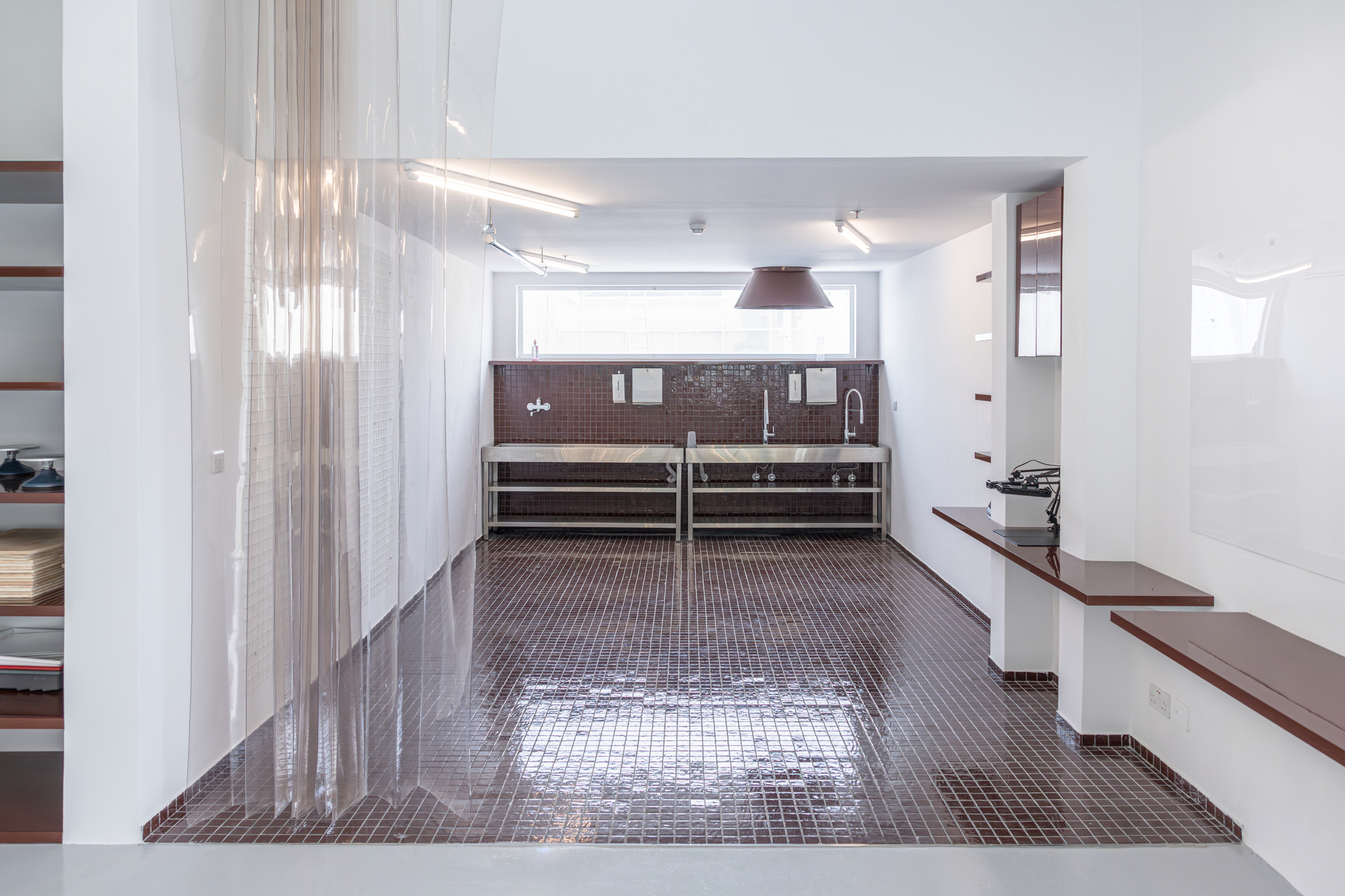 Maroon mosaic-tiled floor and sink wall seen past a hanging strip curtain, with a conical pendant lamp and a window of daylight
