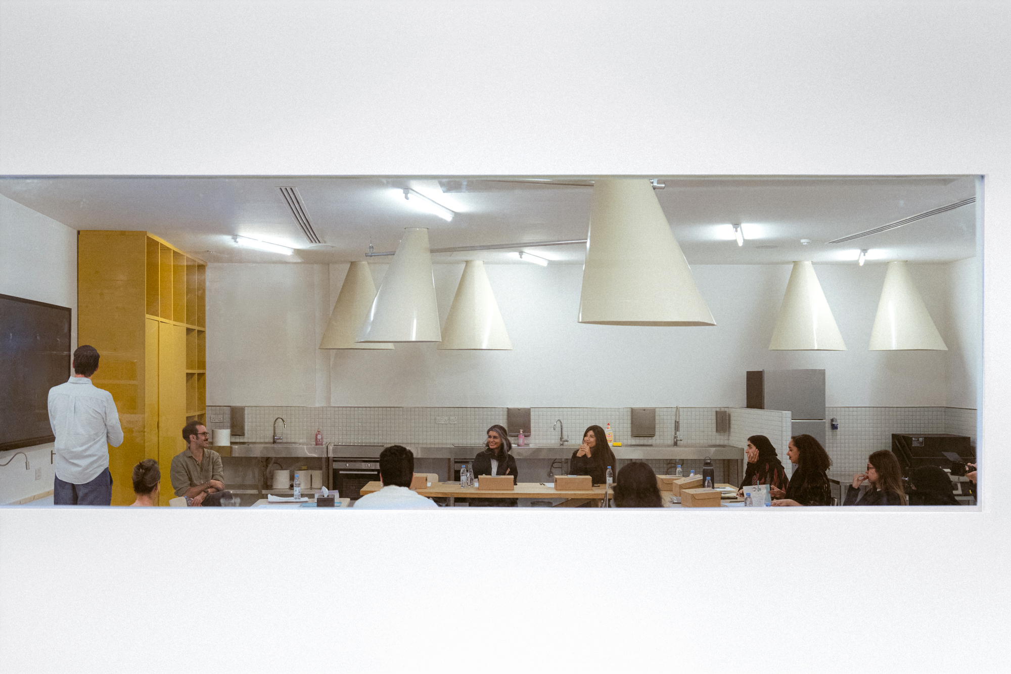 Seated group and a standing speaker viewed through a framed window, under white conical pendant lamps beside a yellow cabinet