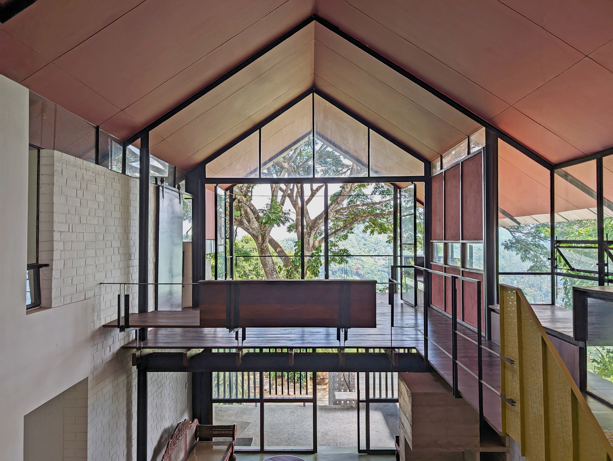 Interior of the gabled double-height space with steel-framed glazing, a timber bridge and a large tree filling the view