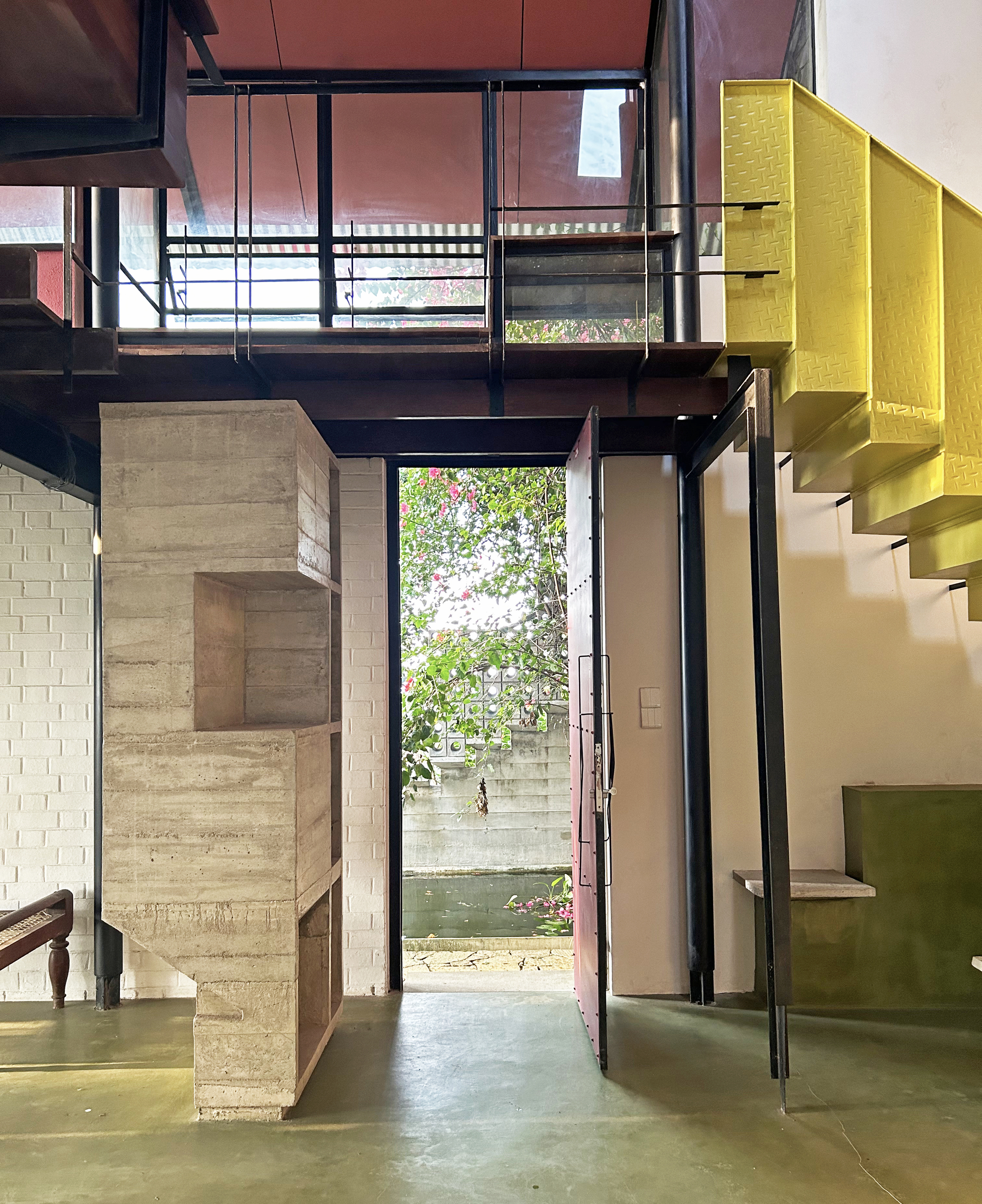 Entry hall with a board-marked concrete block, open pink pivot door and a yellow perforated steel stair above a green floor