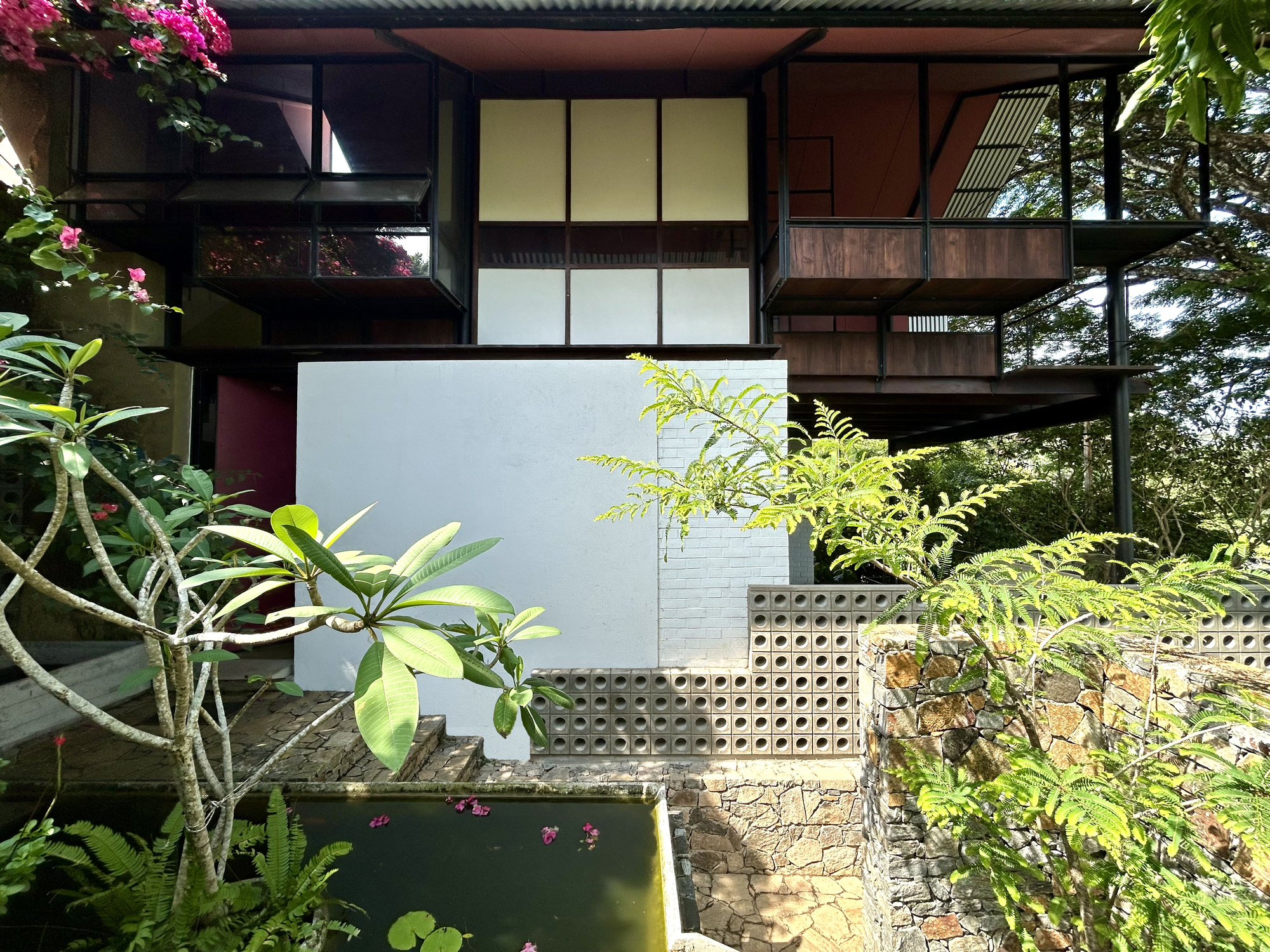 Stepped facade of white wall, timber balconies and perforated block screen above a lily pond, framed by frangipani and bougainvillea