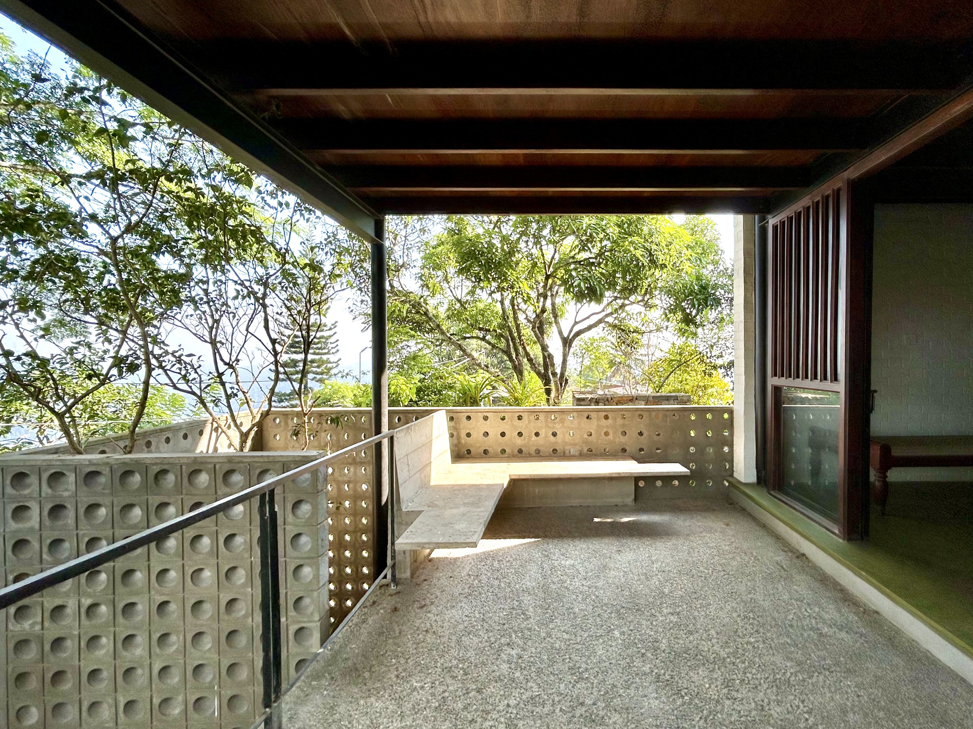 Covered terrace with a built-in concrete bench, perforated block walls and exposed timber beams, framing treetops in daylight