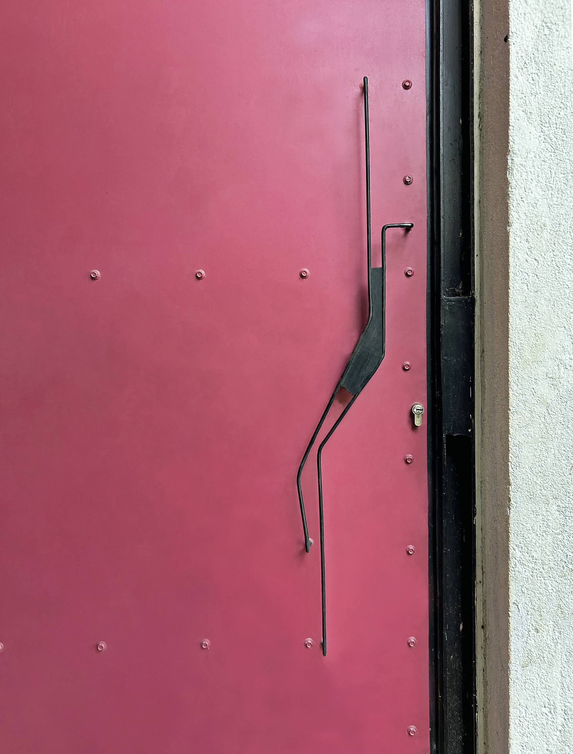 Close-up of a crimson steel door with exposed rivets, a bent wire handle and a cylinder lock beside a white plastered wall