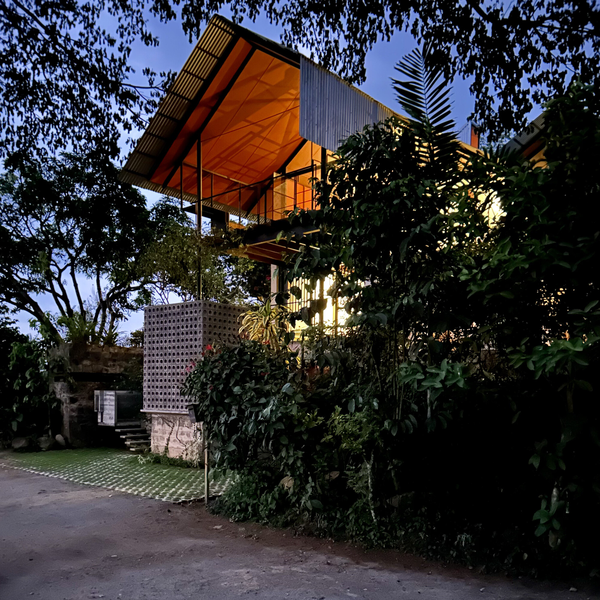 Dusk view of a lit orange-lined timber roof above a perforated block wall, framed by dense foliage and overhanging branches