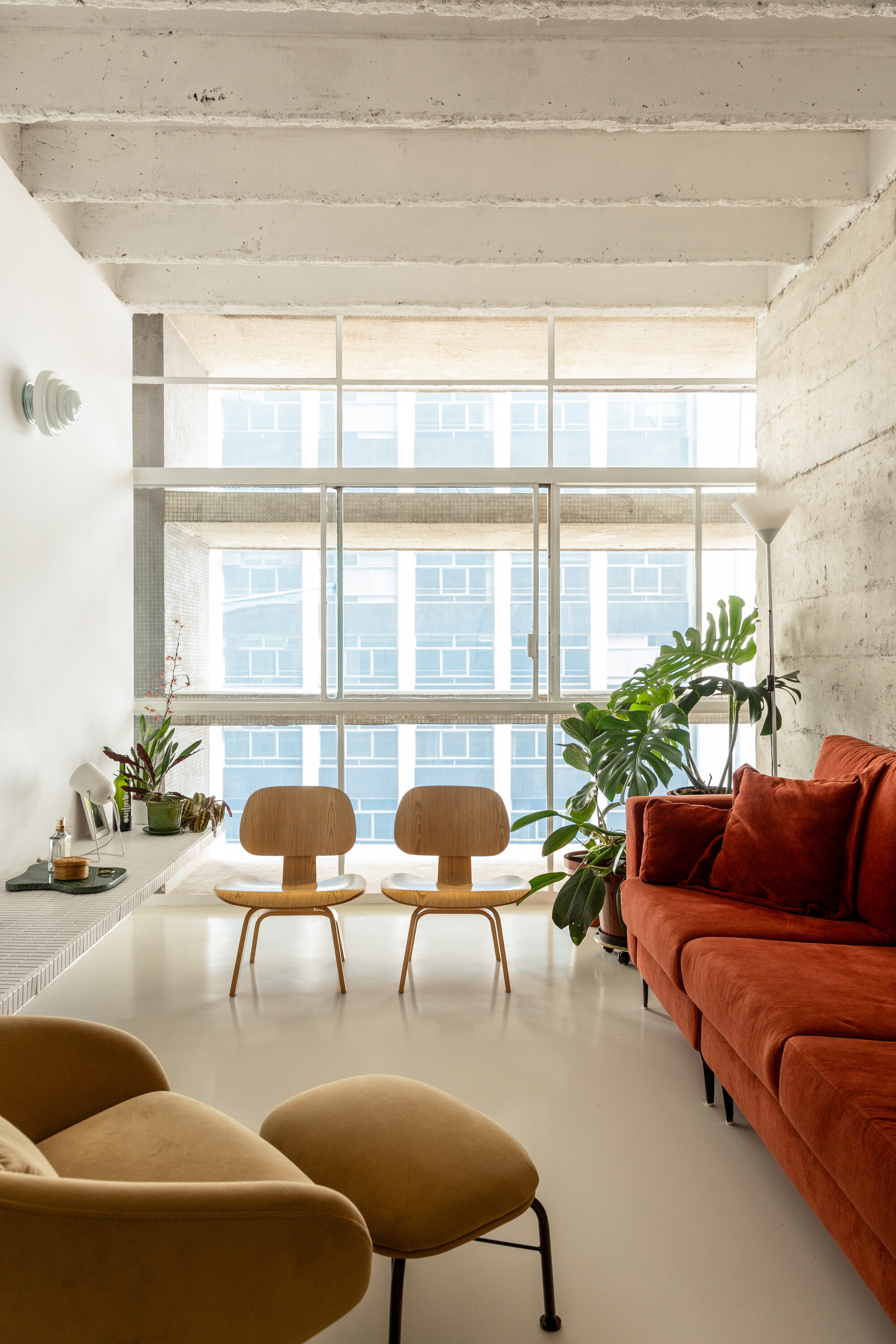 Two plywood lounge chairs face a gridded window wall, with a rust sofa and monstera plant beside exposed concrete beams