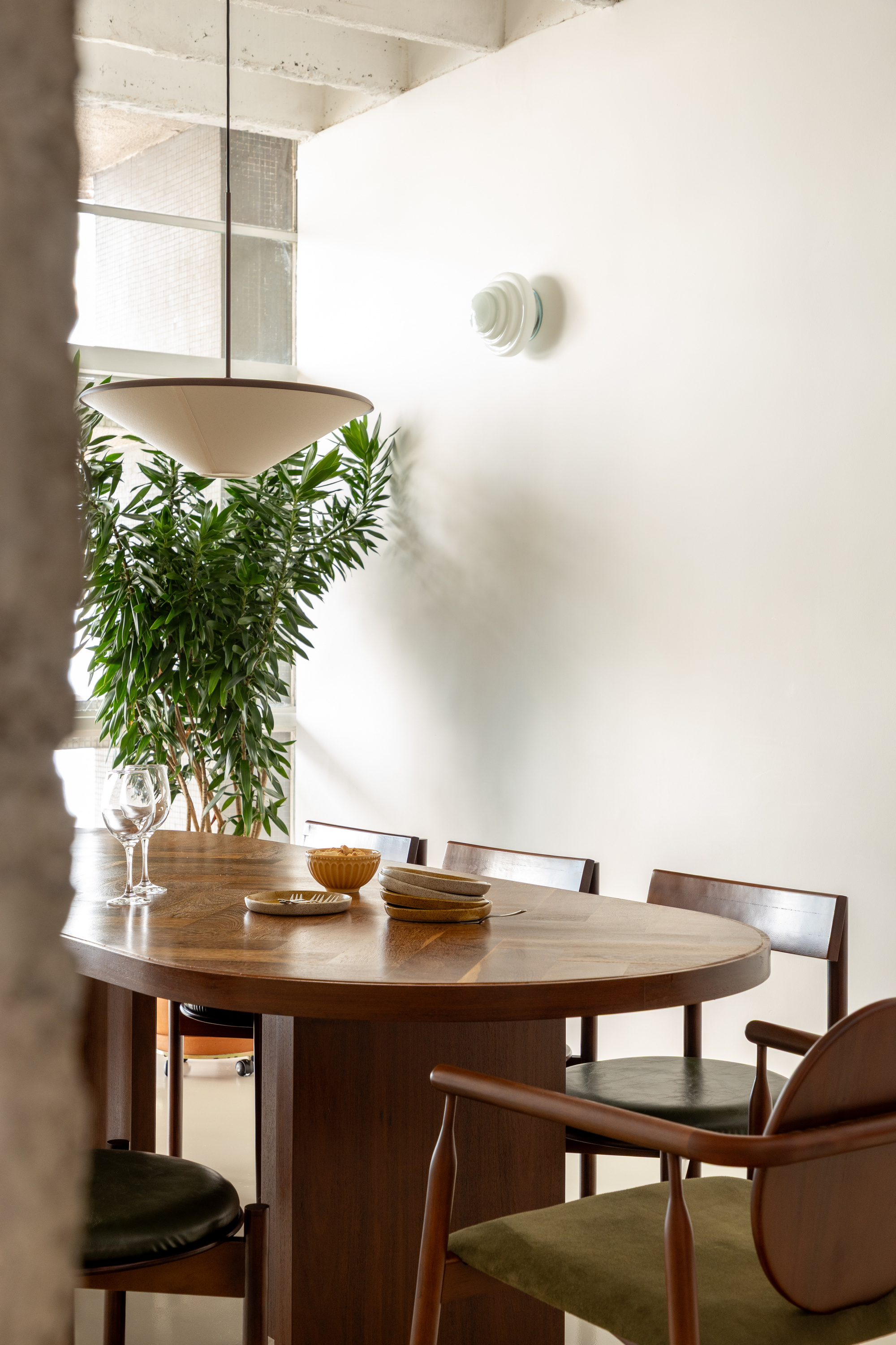 Oval walnut dining table with green upholstered chairs and a conical pendant lamp beside a tall potted plant in soft daylight