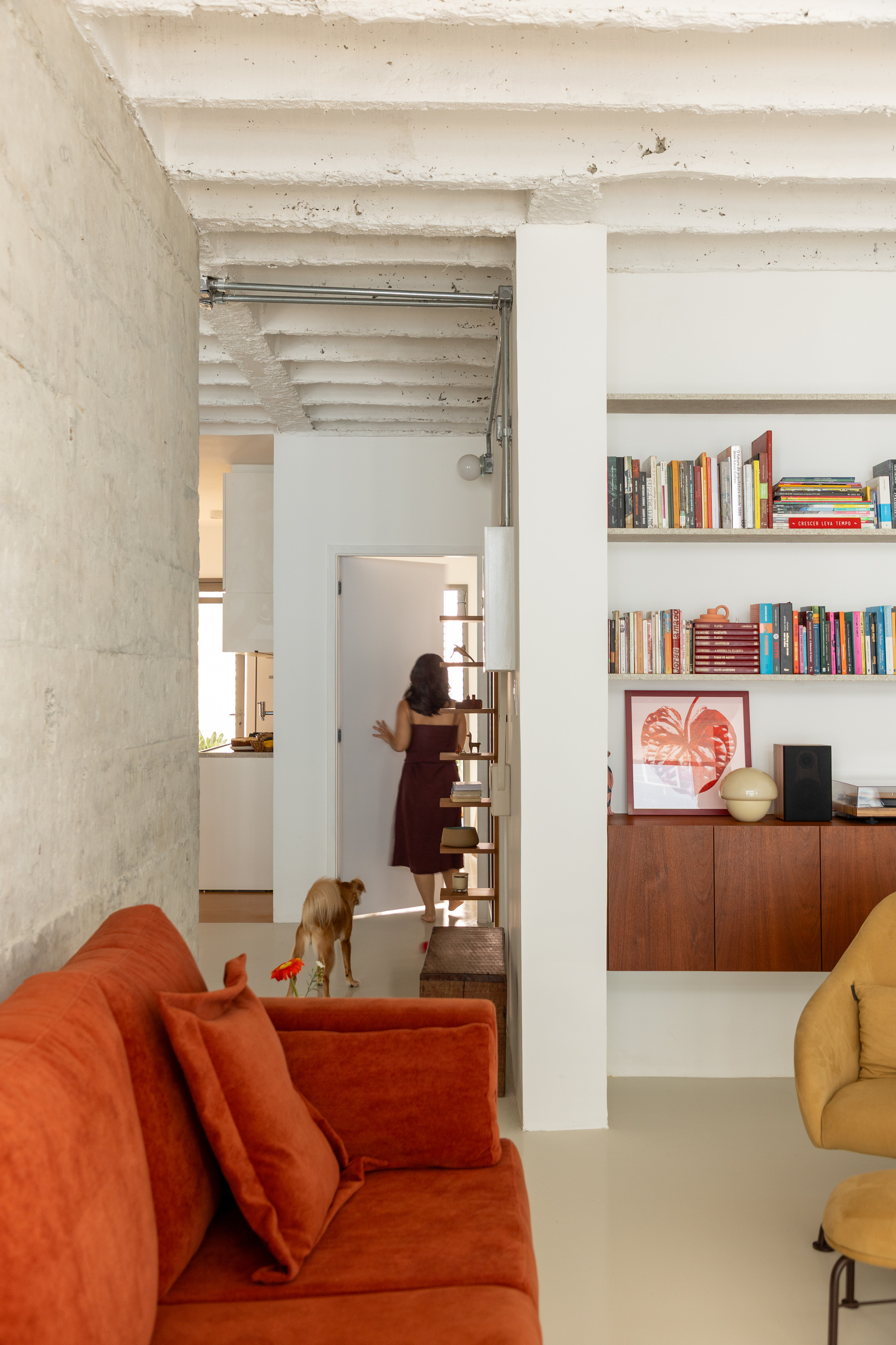 View past a rust sofa toward a hallway, where a woman in a maroon dress walks with a dog beside built-in bookshelves