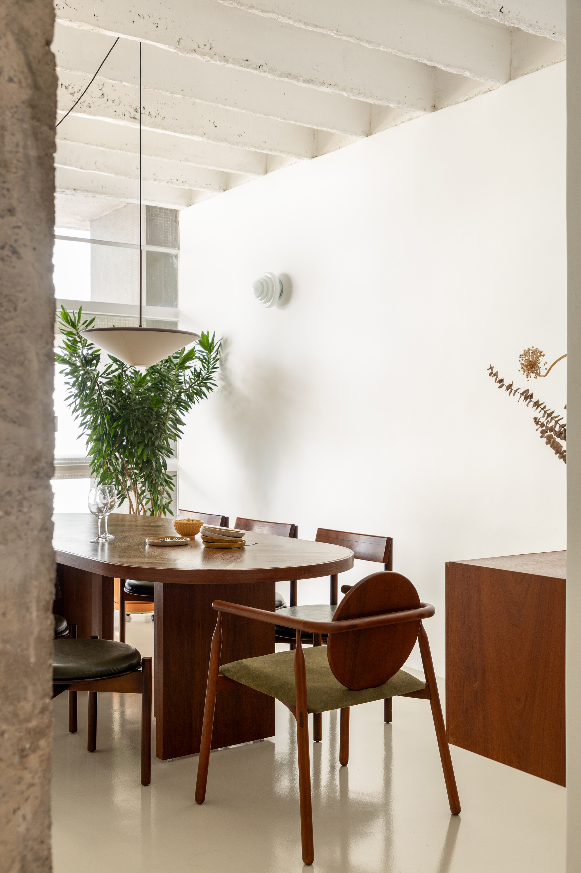 Dining area with an oval walnut table, round-backed chair and pendant lamp beneath white-painted concrete beams, a dried stem at frame edge