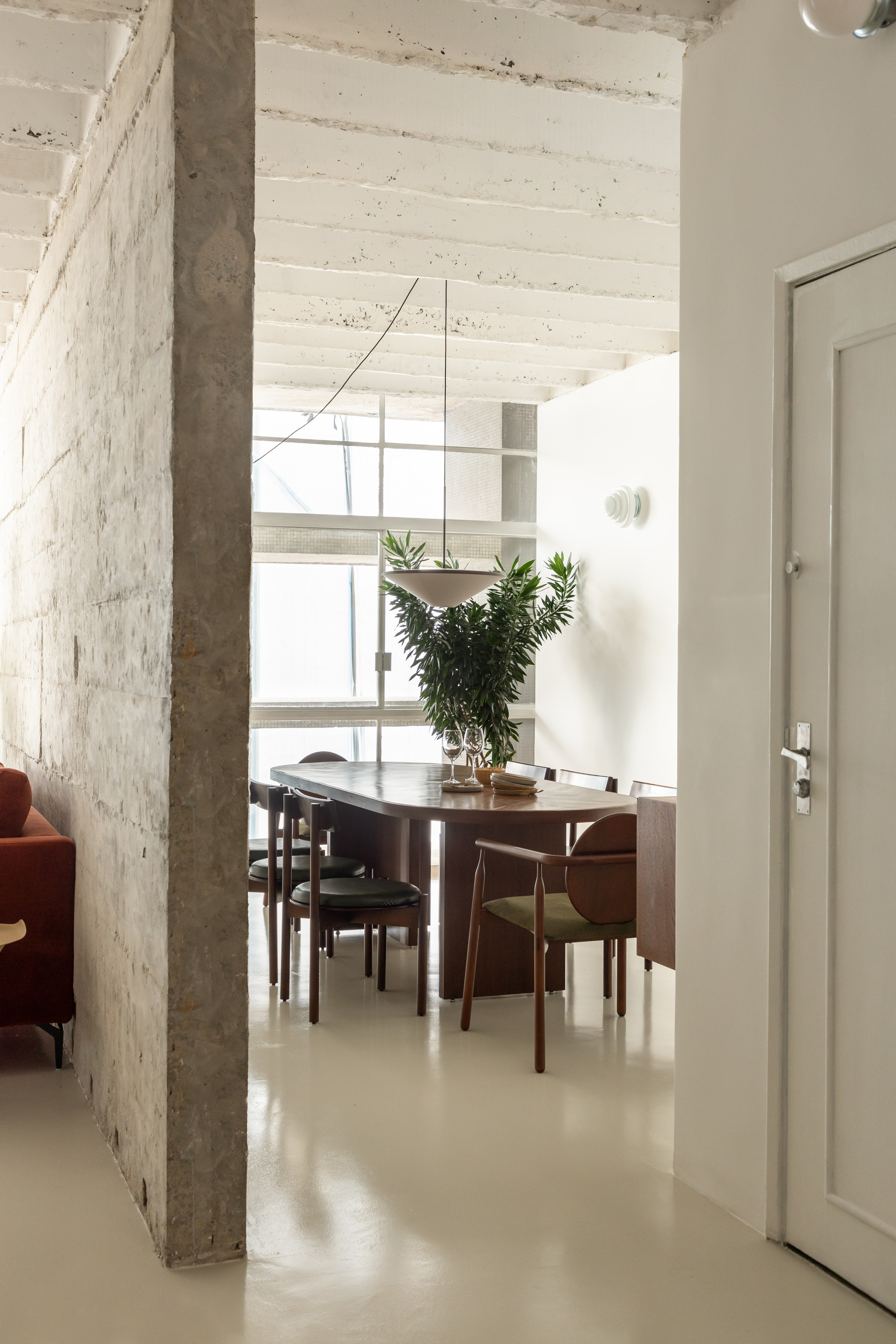 View past a concrete wall into the dining area, with an oval wooden table under a hanging planter and white beams