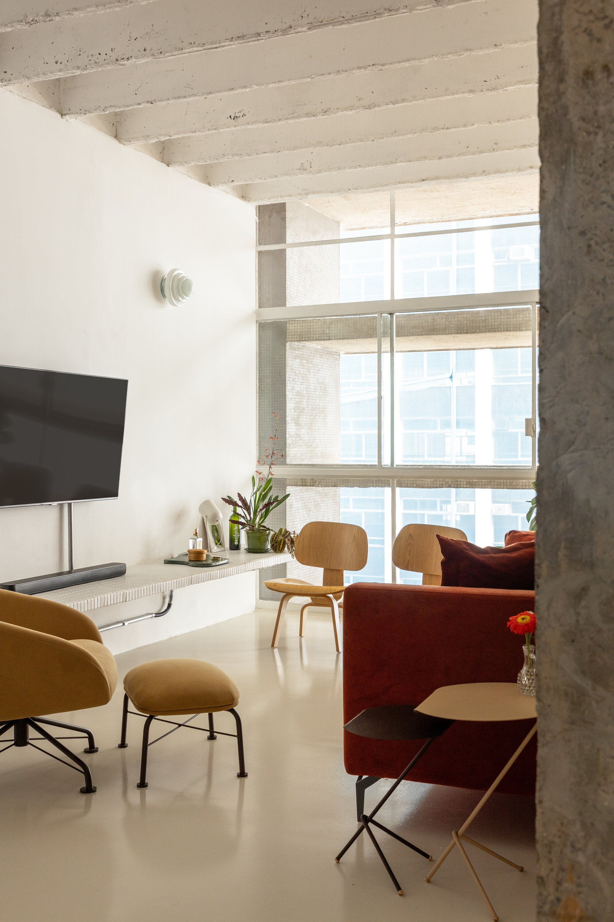 Living area with a rust velvet sofa, plywood lounge chairs and a wall-mounted television, lit by a tall window