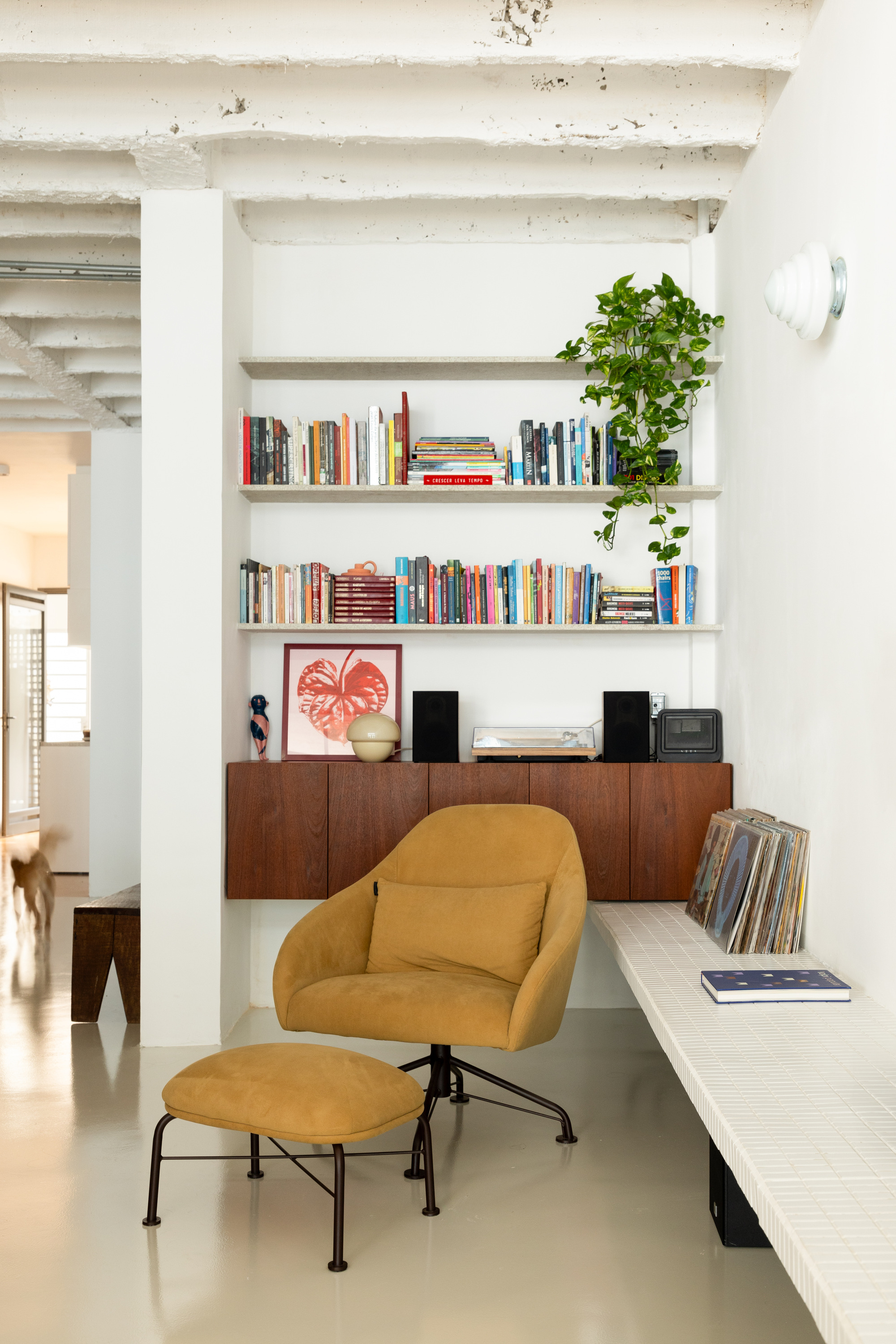 Mustard armchair and ottoman in front of built-in bookshelves, a walnut cabinet and a tiled bench holding records, with a dog blurred at left