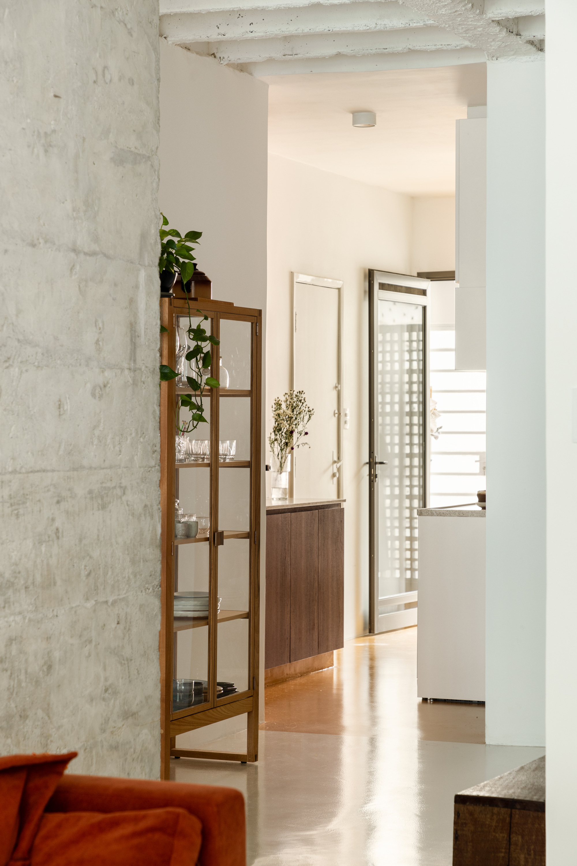 Glass-fronted wooden display cabinet beside a concrete column, with a corridor leading to a perforated door in soft daylight