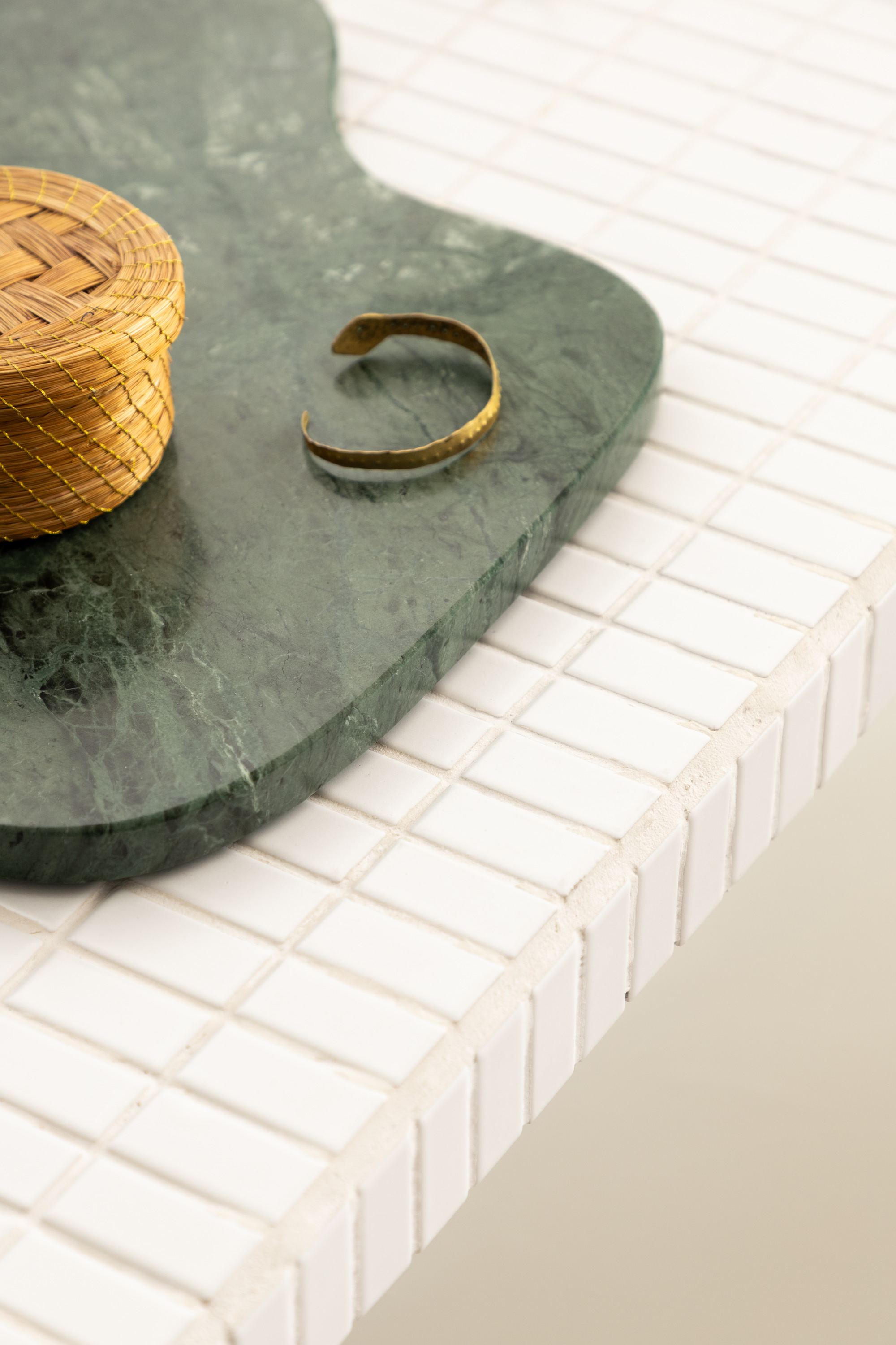 Corner of a white mosaic tile tabletop holding a green marble tray, a woven straw box and a brass cuff in soft light