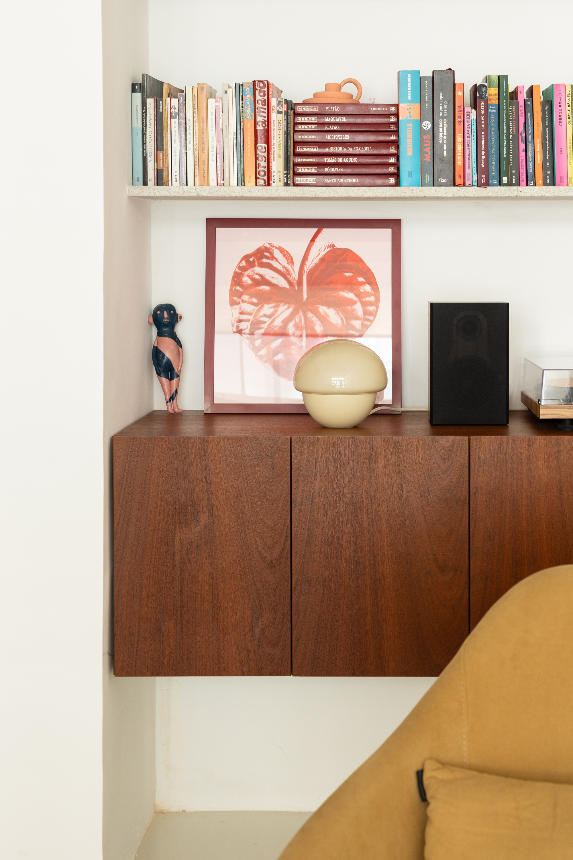 Floating teak cabinet in a white niche with a framed anthurium print, a cream lamp, a speaker and books above