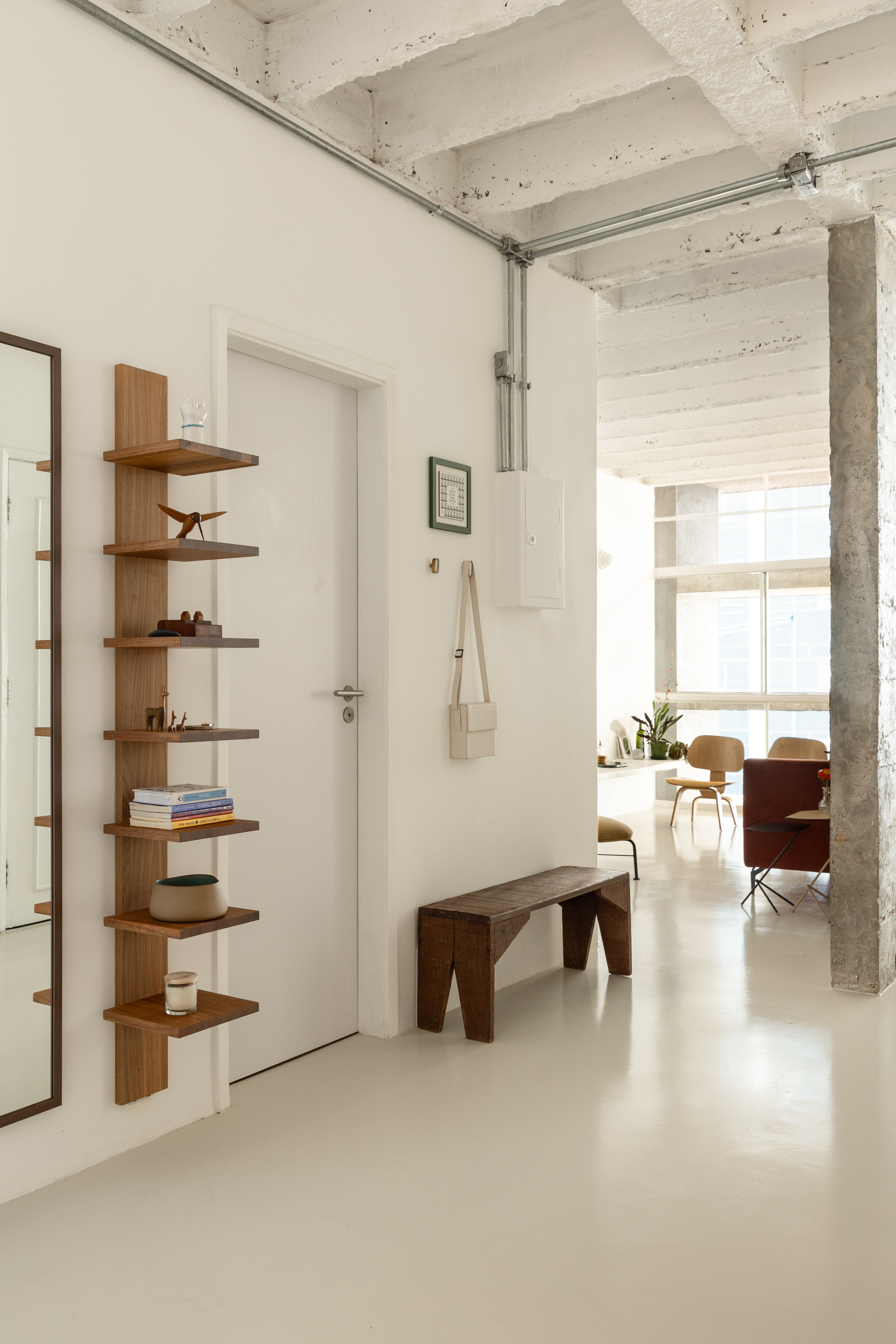 Entry hall with a wall-mounted timber shelf column, a rough wooden bench and exposed painted concrete beams in soft daylight