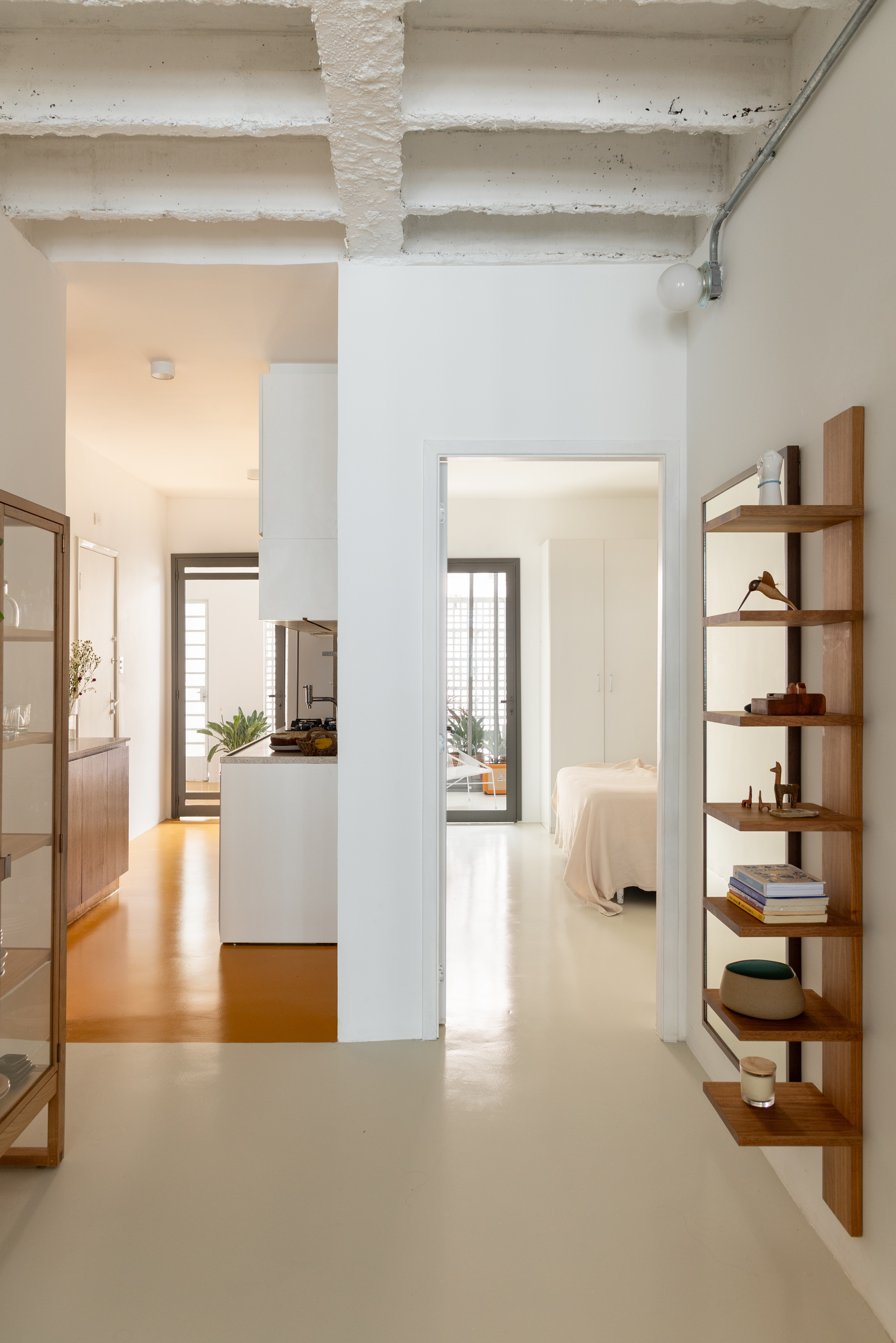 View down a pale resin-floored corridor toward a kitchen with an ochre floor and a bedroom, under white-painted concrete beams